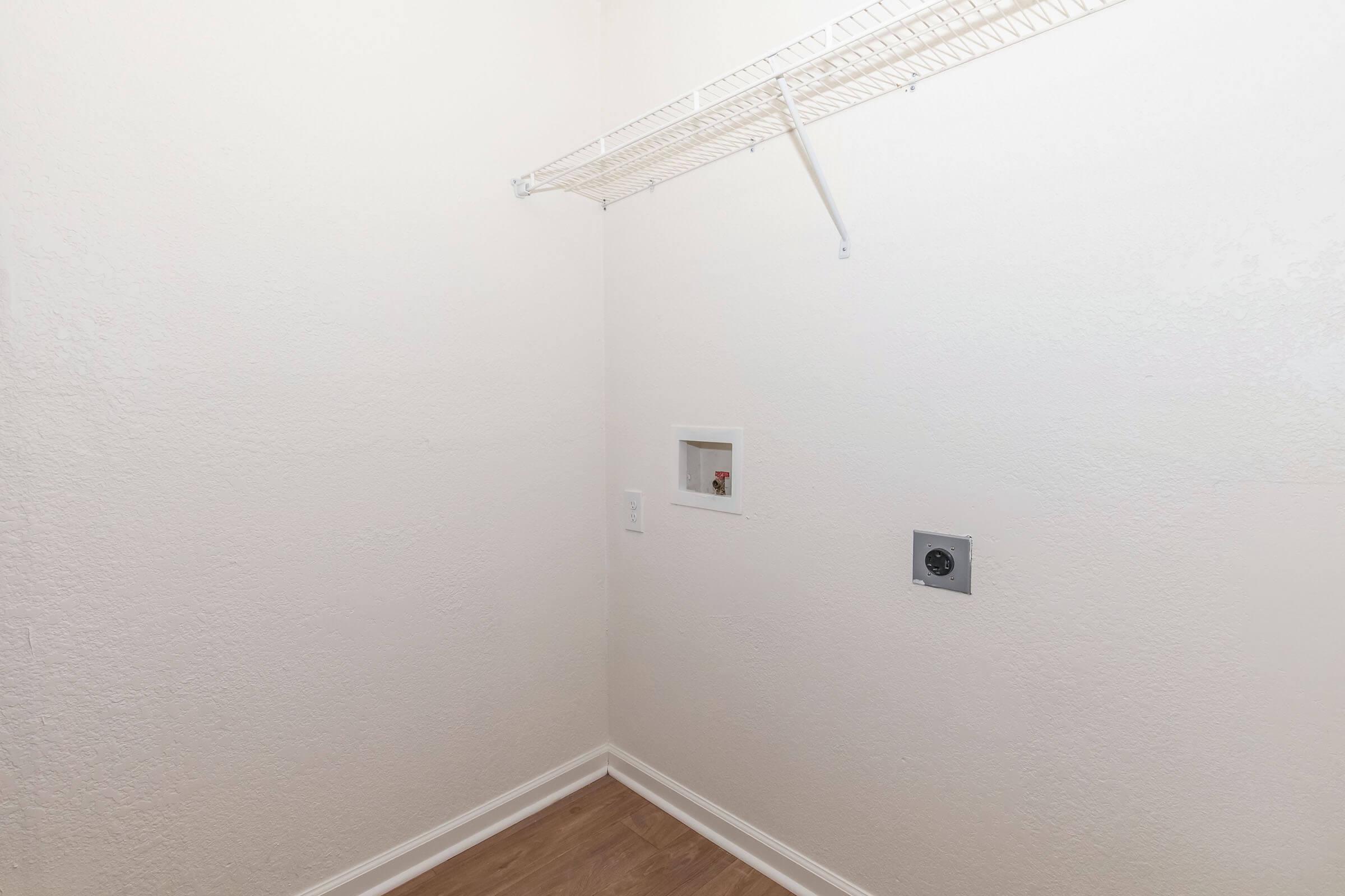 A simple, empty laundry room featuring light-colored walls, a wire shelf mounted on the wall, and a small opening in the wall likely for plumbing. The floor is wooden, and there's an electrical outlet visible on the wall. Overall, the space appears clean and ready for appliances.