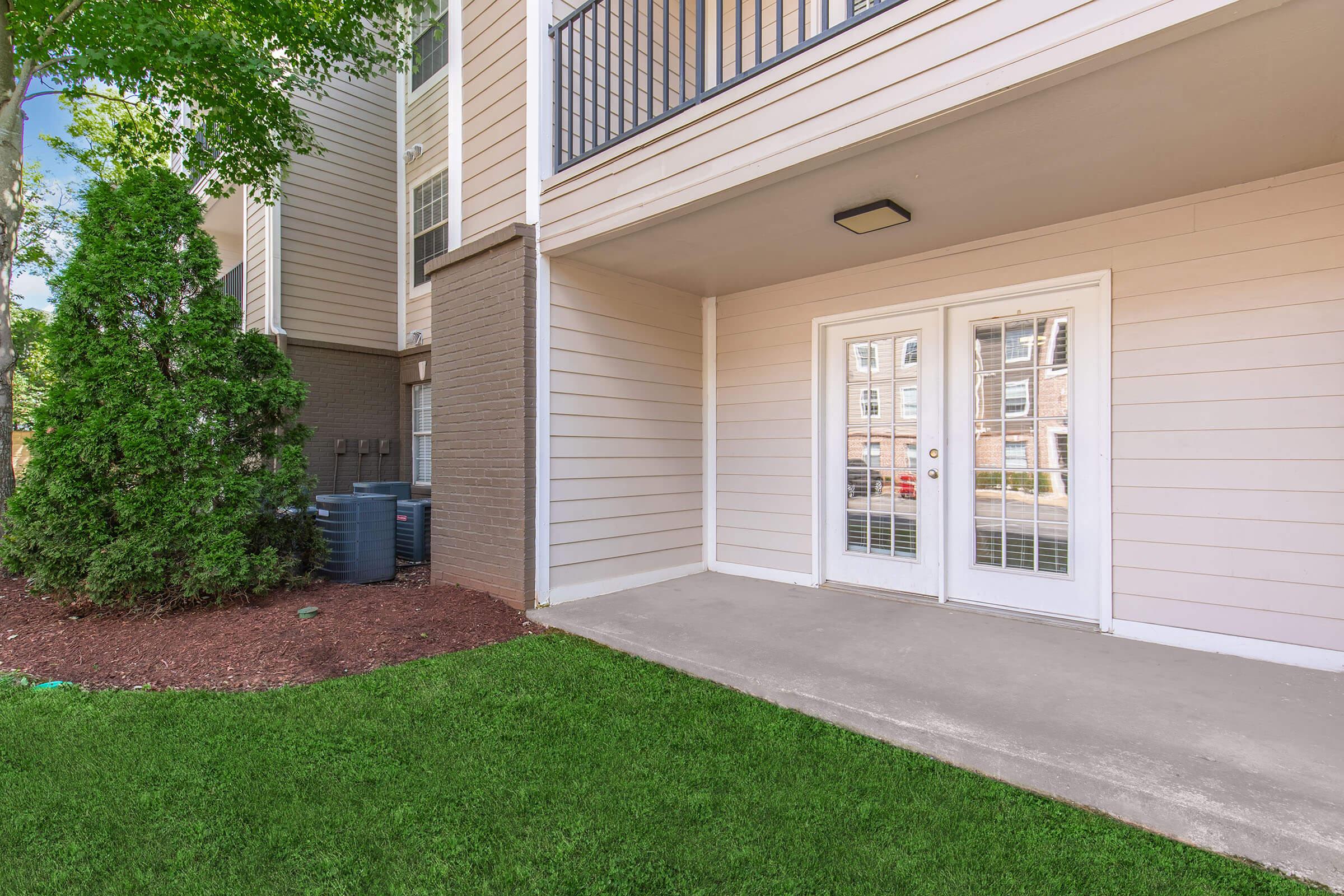 A view of an apartment exterior featuring a small patio area with double glass doors, surrounded by green grass and shrubs. The building has light-colored siding and a balcony above. In the background, utility boxes can be seen beside the building.