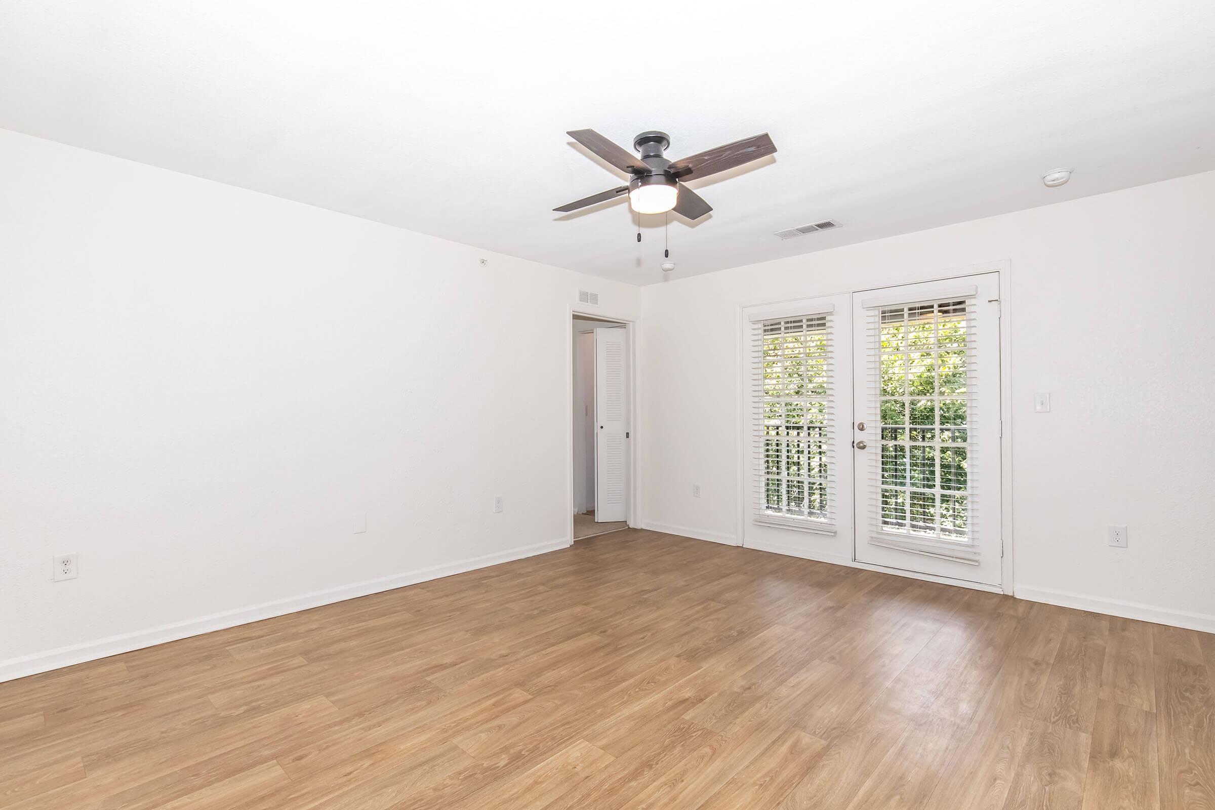 A spacious, empty room featuring light wood flooring, white walls, and a ceiling fan. Large windows with shutters allow natural light to brighten the space. A door leads to another area, enhancing the openness of the room. The overall ambiance is bright and inviting, perfect for personalized decoration.