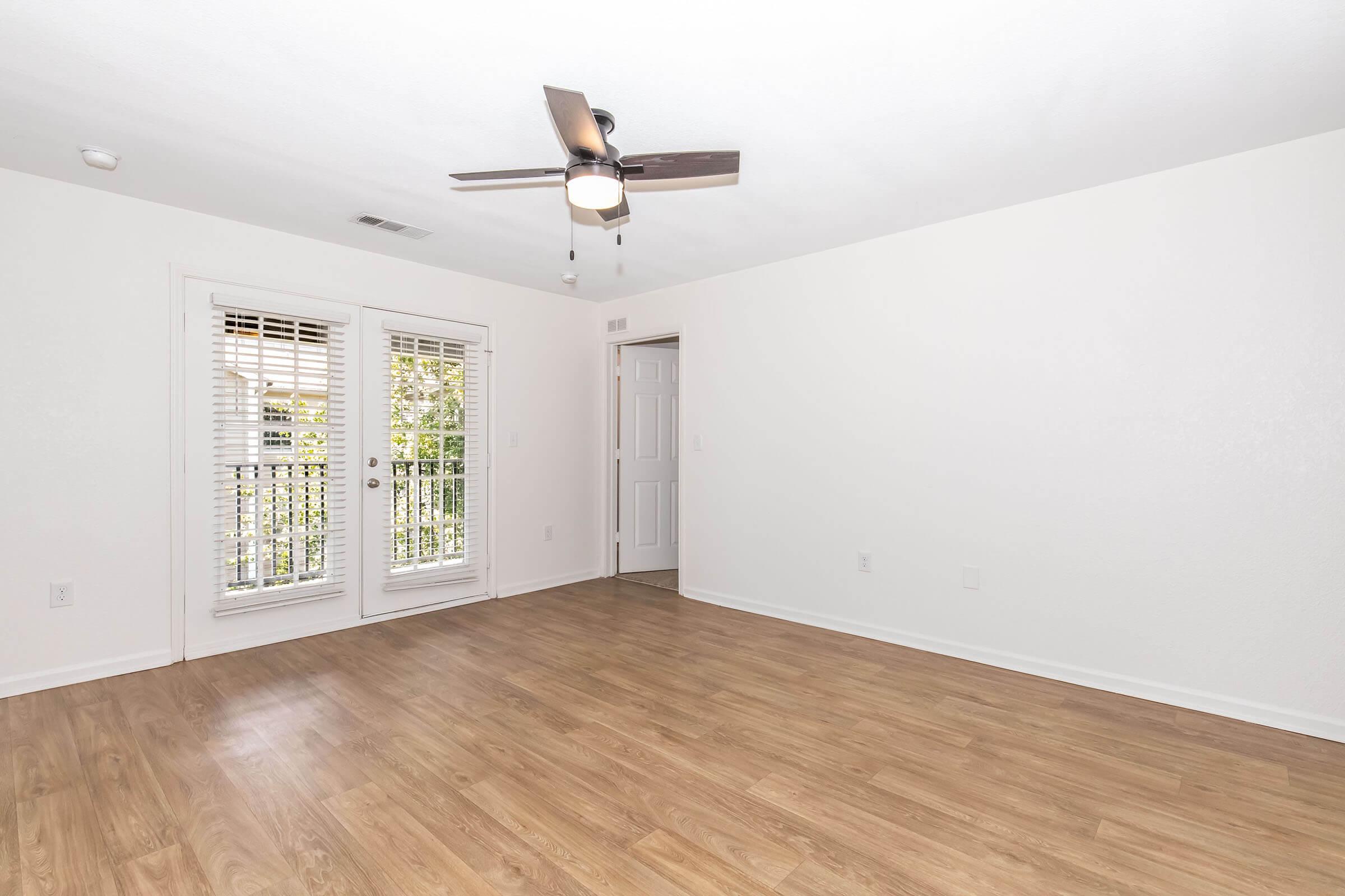 Empty room with light-colored walls and wood-style flooring. A ceiling fan is mounted in the center. There are two large windows with blinds on one side, leading to a small balcony. A white door is visible on the opposite wall. The space is bright and airy, creating an inviting atmosphere.