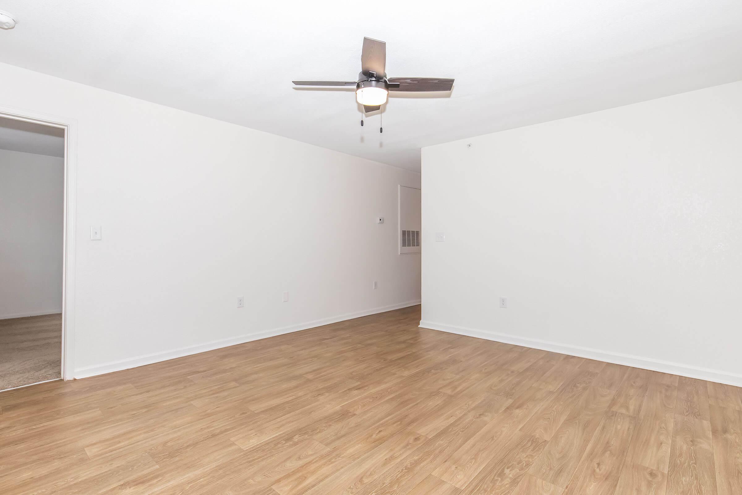 A spacious, empty room with light-colored walls and laminate flooring. There is a ceiling fan with light fixtures. One wall features a doorway leading to another area, while a small window can be seen in the background. The room has an inviting, well-lit atmosphere.