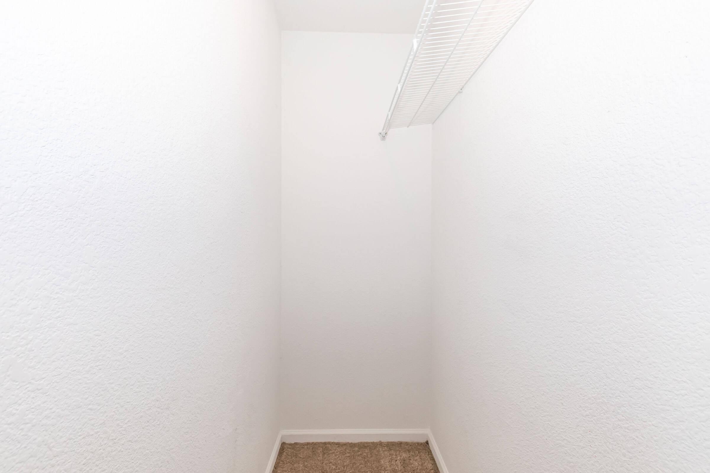 Empty closet with white walls and a beige carpeted floor. A wire shelving unit is mounted along the top wall, providing extra storage space. The overall space is well-lit and reflects a minimalist design. No items are present in the closet.