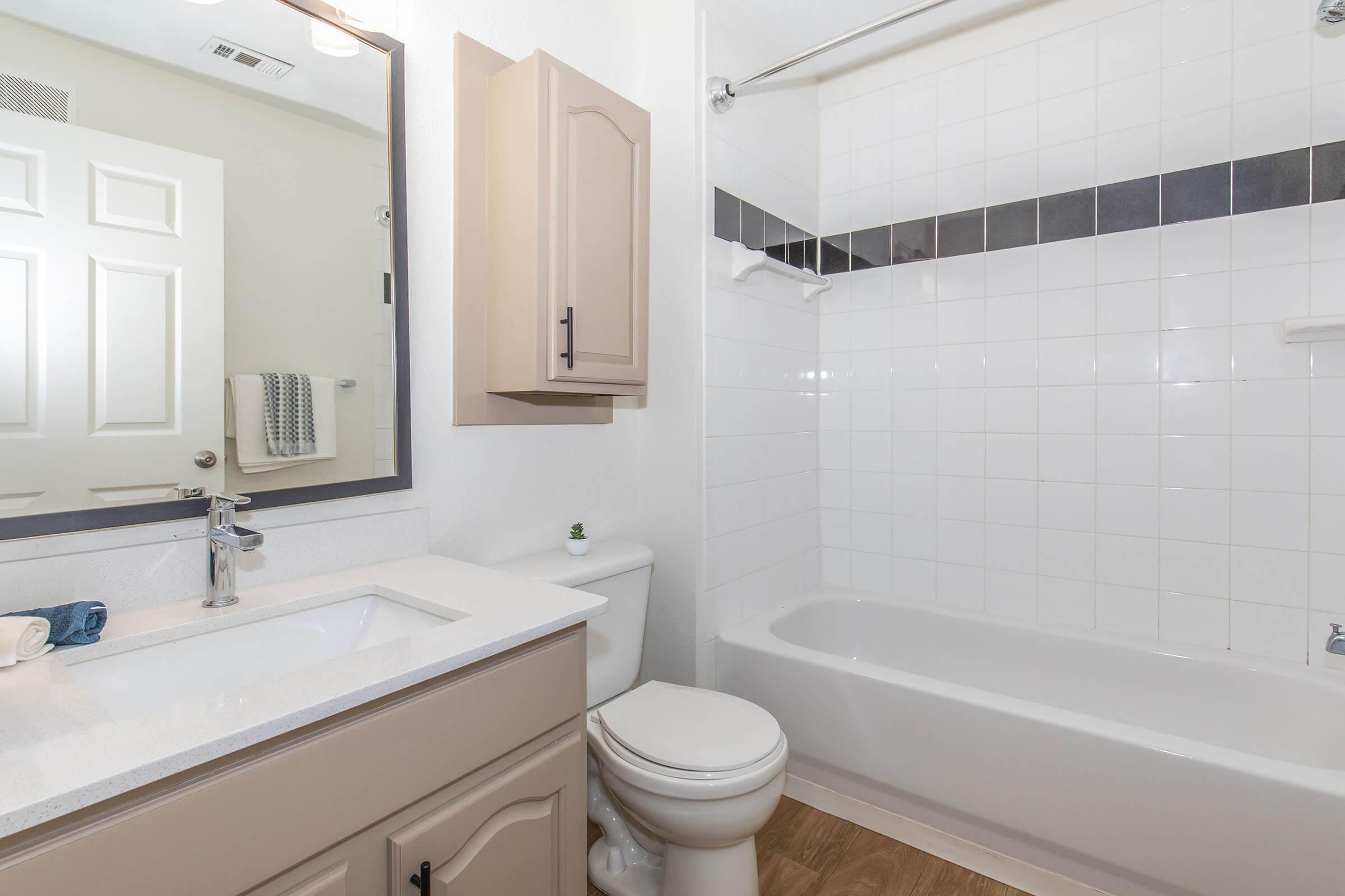 A clean and modern bathroom featuring a bathtub, a sink with a vanity, and a large mirror. The walls are tiled in white with a decorative black stripe. There’s a towel rack and a bathroom cupboard, complemented by wood-style flooring. Bright lighting enhances the airy feel of the space.