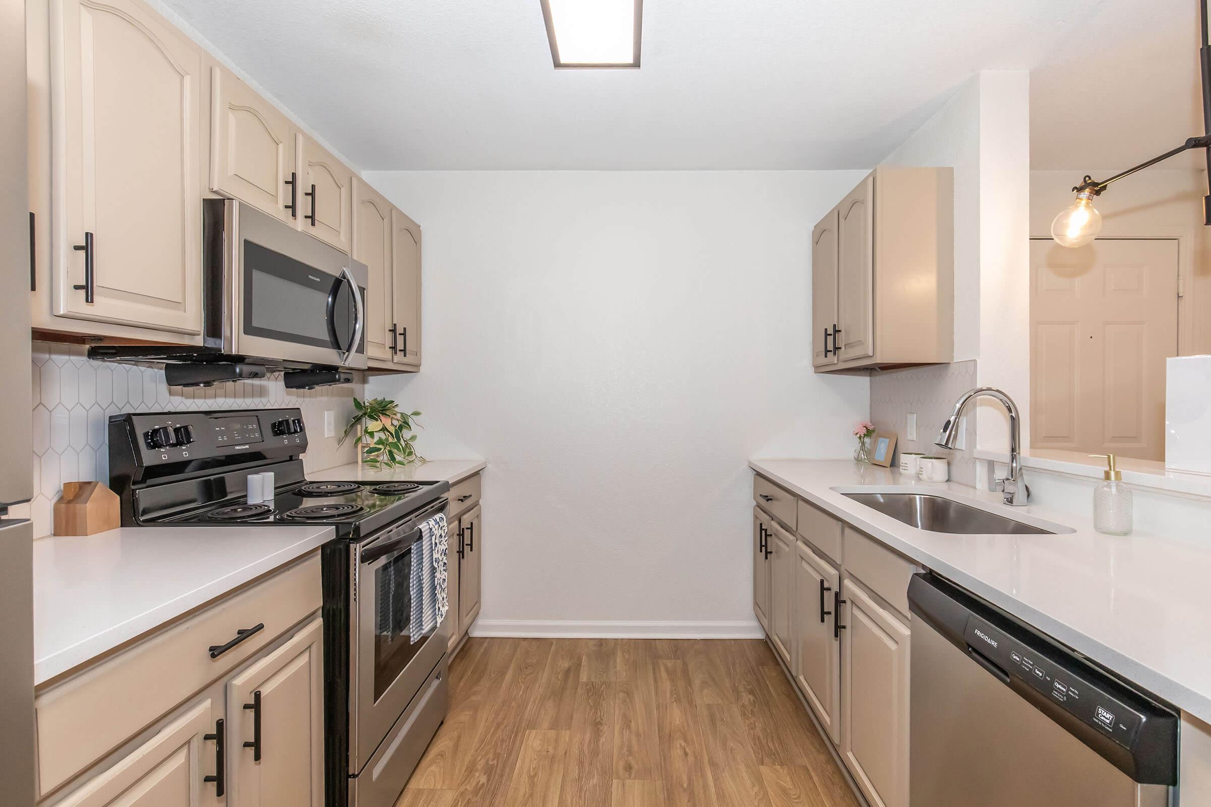 Modern kitchen featuring light beige cabinetry, stainless steel appliances including a stove and microwave, and a white countertop. The kitchen includes a double sink with a sleek faucet, and a decorative plant on the countertop. The flooring is light wood, contributing to a bright and airy atmosphere.