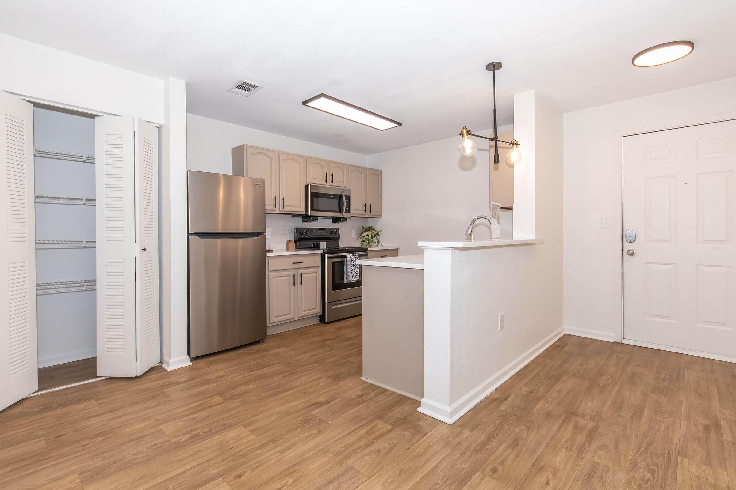 Modern kitchen and dining area featuring stainless steel appliances, including a refrigerator and stove. Light wood flooring throughout enhances the bright, open space. A pantry is visible with sliding doors, and there is a welcoming entryway with a front door. Decorative light fixtures add a contemporary touch.