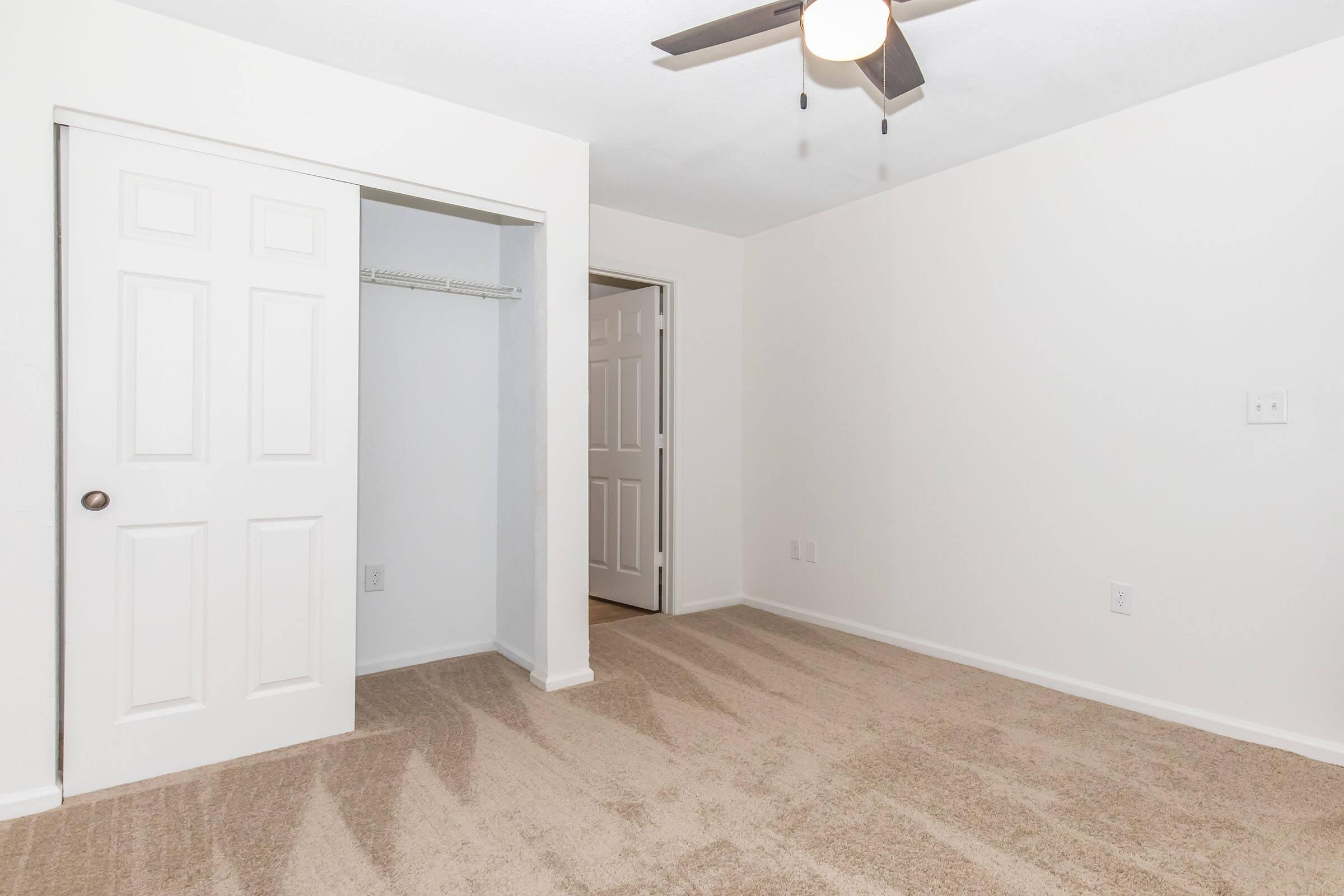 A bright, empty room with beige carpet and white walls. There's a ceiling fan and two closed doors, one leading to a closet and another to an adjoining space. The room appears clean and freshly painted, suitable for various uses like a bedroom or office.