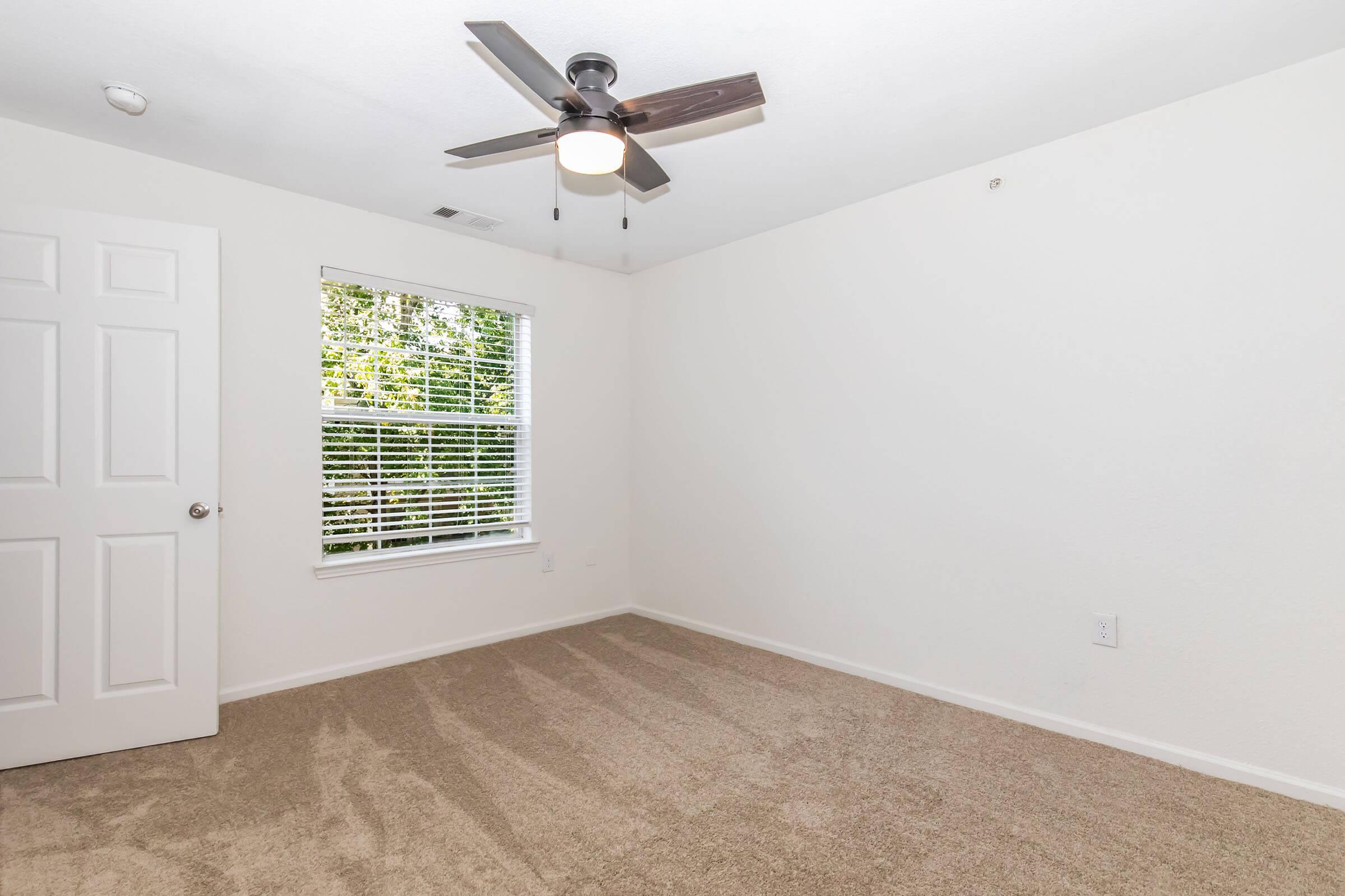 Empty room featuring light-colored walls, a ceiling fan with a light fixture, and a window with blinds allowing natural light. The floor is carpeted in a neutral tone. A closet door is visible on the left side of the room.