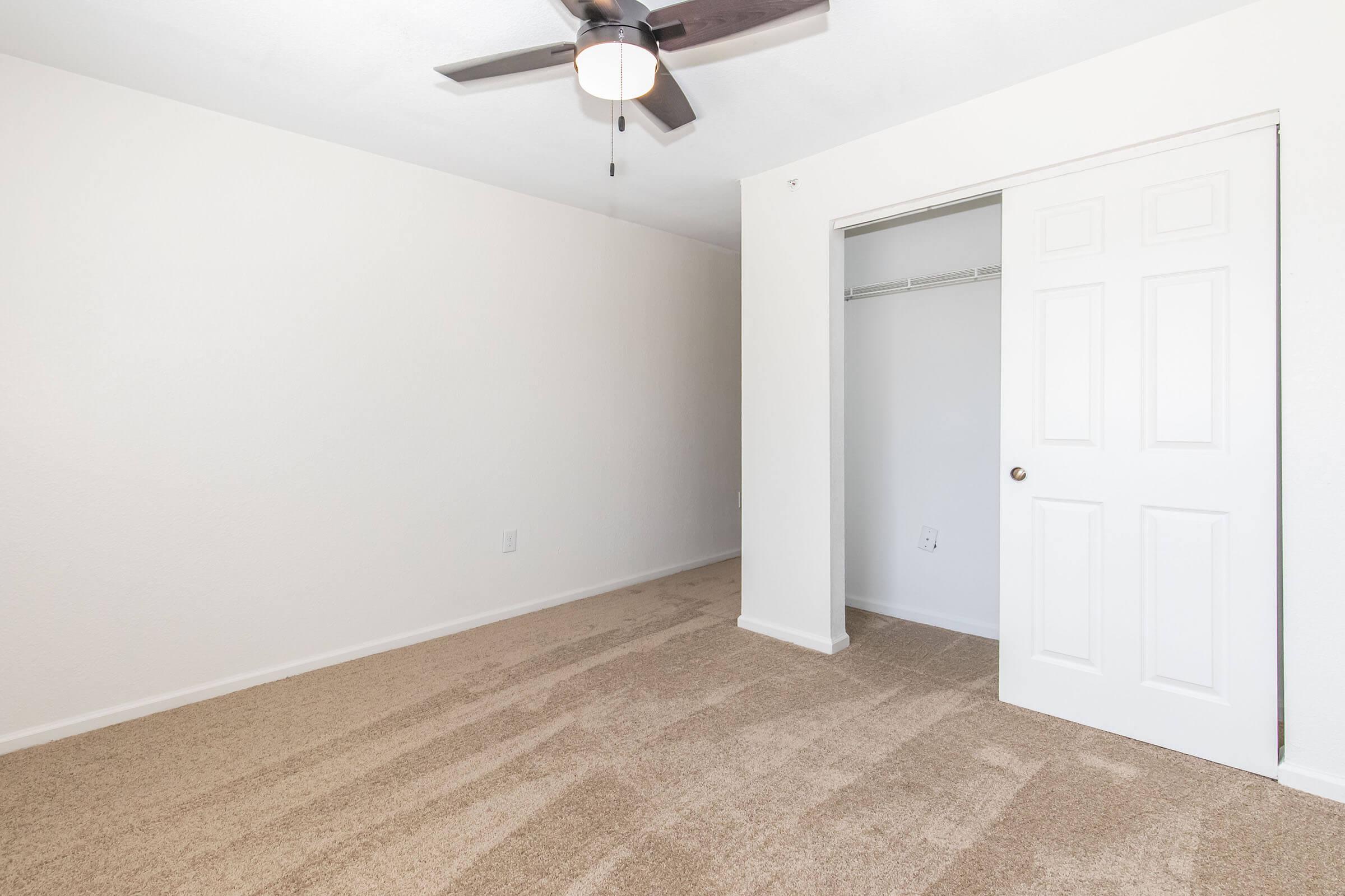 A clean, empty room with light beige carpet and white walls. There is a ceiling fan installed, and a closet with sliding doors is visible. Natural light fills the space, creating a bright atmosphere. The room appears freshly painted and well-maintained.