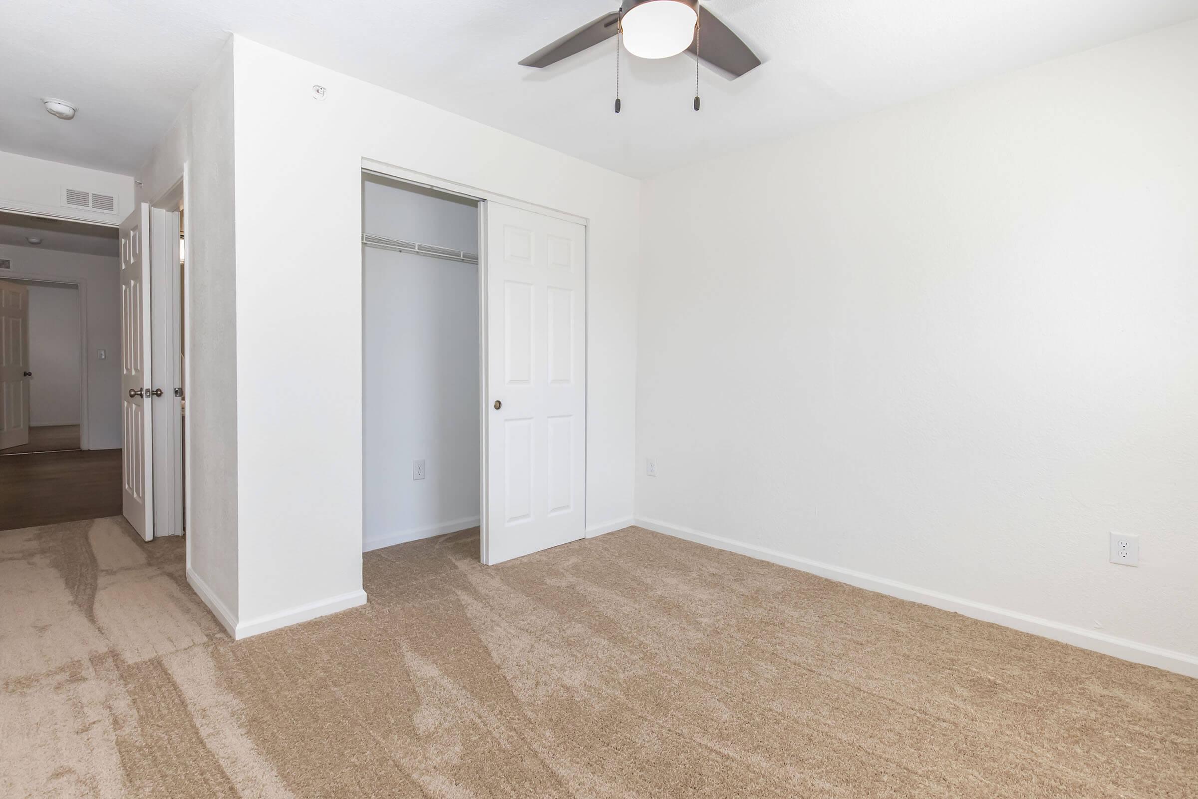 A spacious, empty room with beige carpet and white walls. There is a ceiling fan installed, and a closet with double doors is visible on the left side of the image. The room is well-lit, showcasing ample natural light, with another doorway leading to a different area in the background.