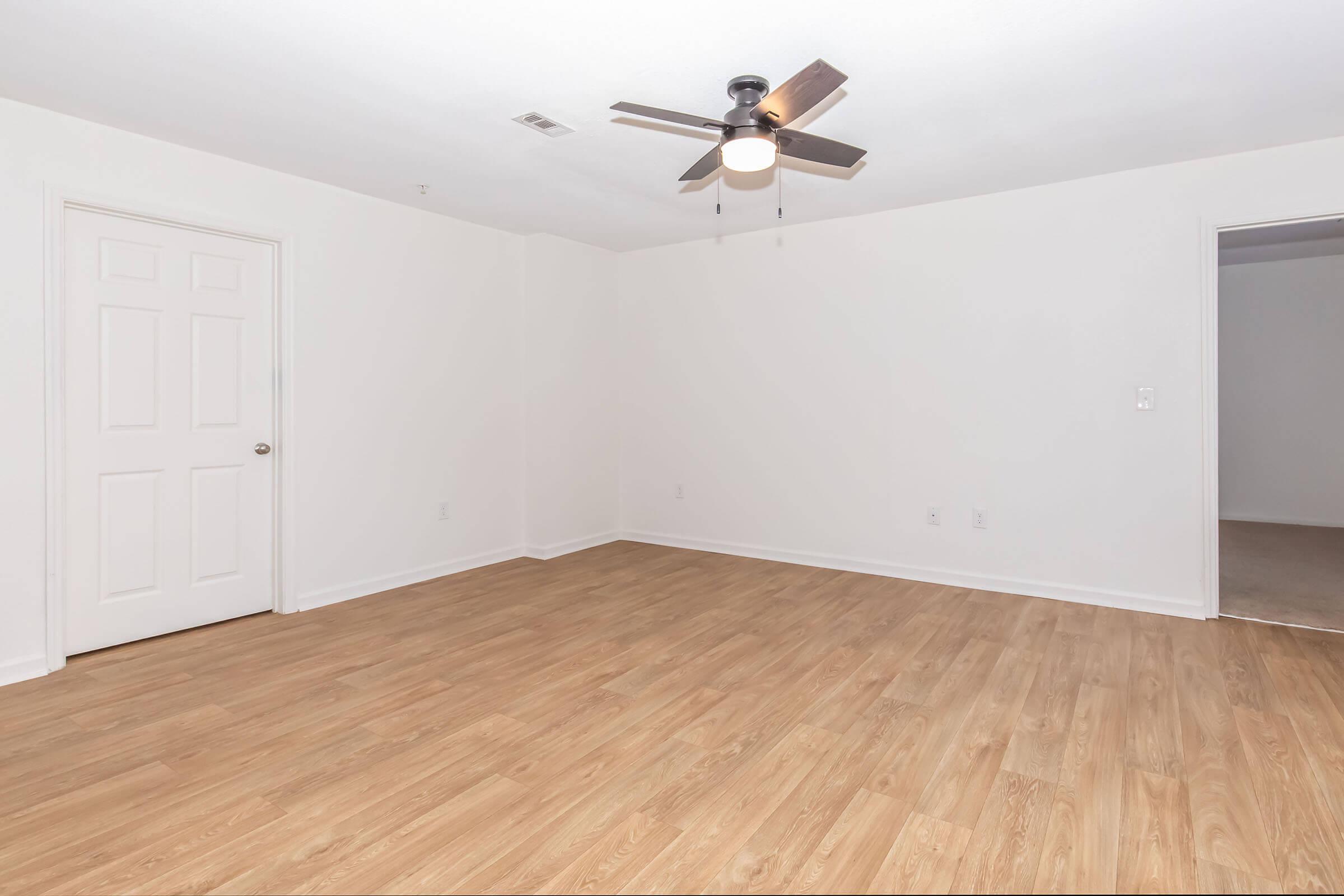 An empty room with light-colored walls and a wooden floor. There's a ceiling fan with light in the center of the ceiling. A closed door is visible on the left, and another room entrance is seen on the right. The space is bright and uncluttered, indicating a fresh or newly renovated area.