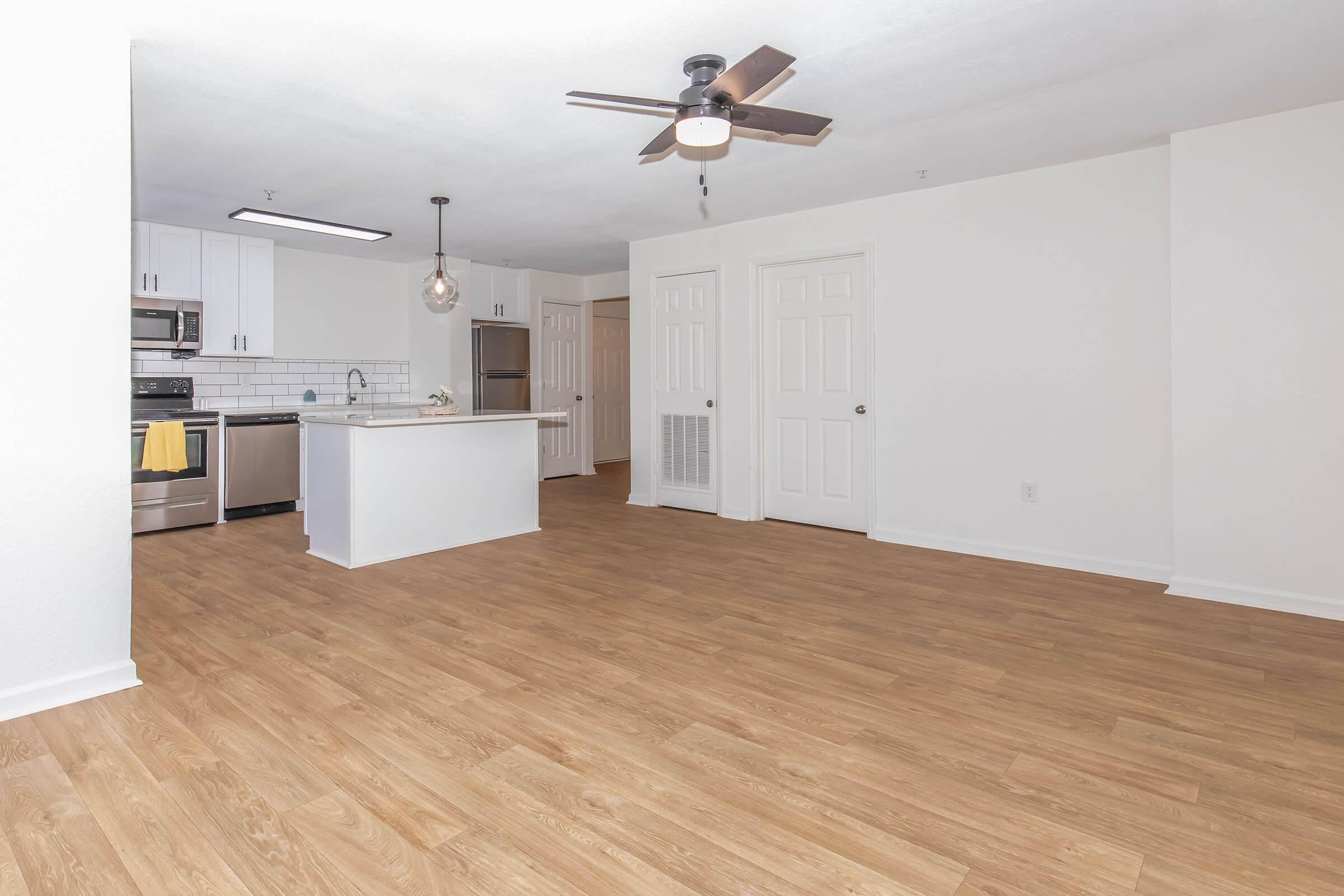 A spacious, well-lit living area with light-colored walls and wood-style flooring. It features an open kitchen with stainless steel appliances, a kitchen island, and modern lighting. The room has a ceiling fan and ample space for furniture, creating a cozy and inviting atmosphere.