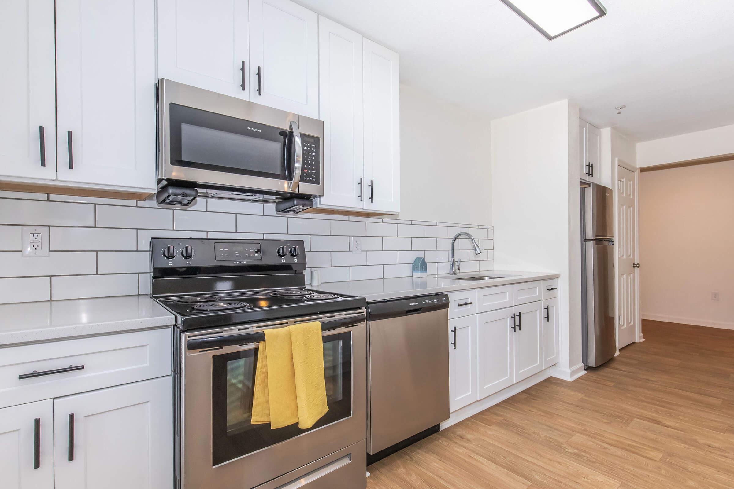Modern kitchen featuring stainless steel appliances, including a microwave, oven, and dishwasher. The kitchen has white cabinets, a light gray backsplash, and a countertop, with a yellow dish towel hanging from the stove. The flooring is light wood, and there is natural light coming from a nearby door.