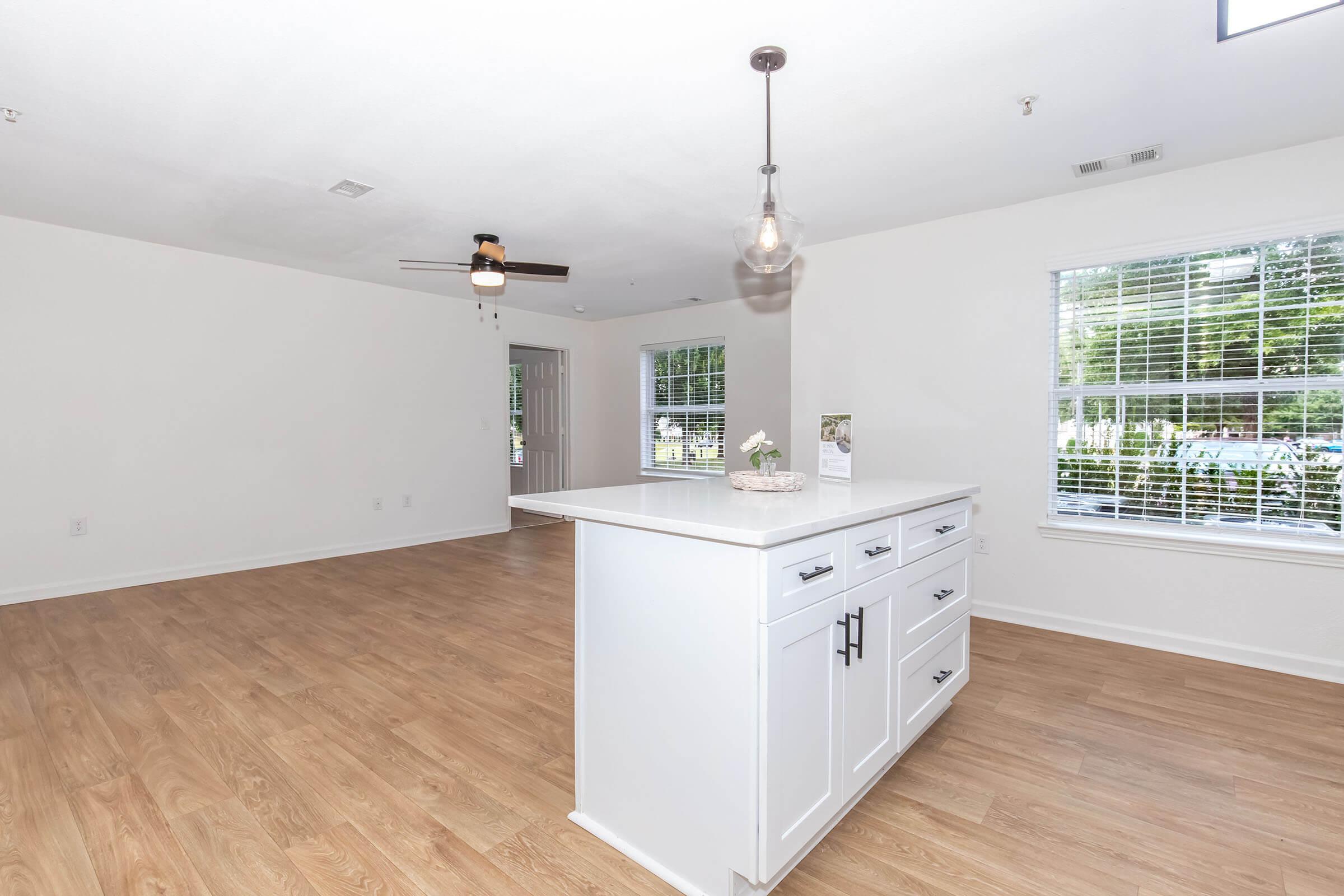 A spacious, modern interior featuring light-colored walls and wood-style flooring. In the foreground, a white kitchen island with drawers and a decorative centerpiece. The background shows a well-lit living area with a ceiling fan and large windows allowing natural light.