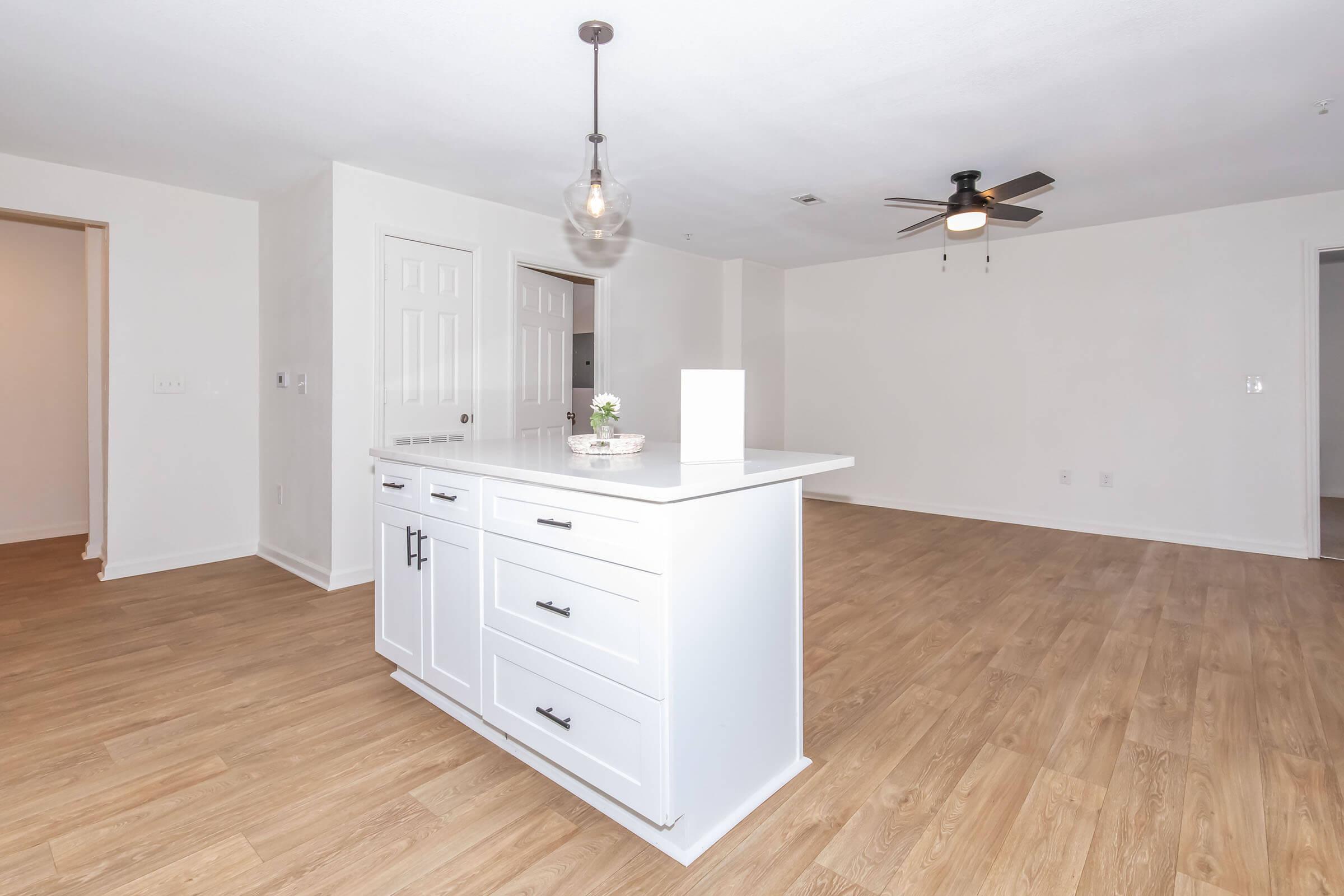 Bright, spacious living area featuring light wood laminate flooring, white walls, and a central kitchen island with modern cabinetry. A contemporary pendant light hangs above the island, while a ceiling fan is mounted on the ceiling. Open concept layout with doorways leading to adjacent rooms.