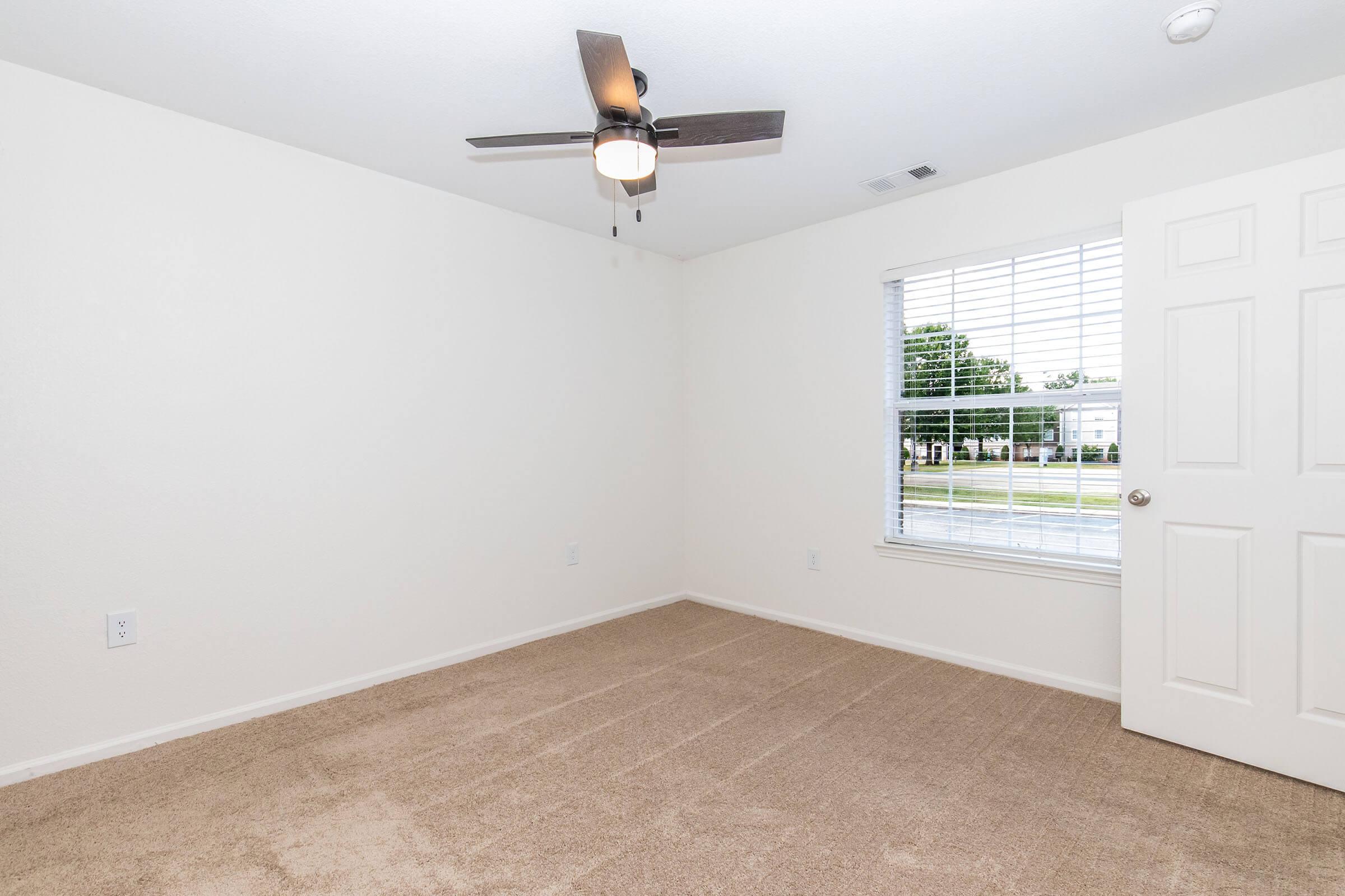 A clean, empty room with light beige carpet and white walls. There's a ceiling fan with light fixtures, a window with blinds letting in natural light, and a closed white door. The room has a simple and spacious feel, suitable for various uses.