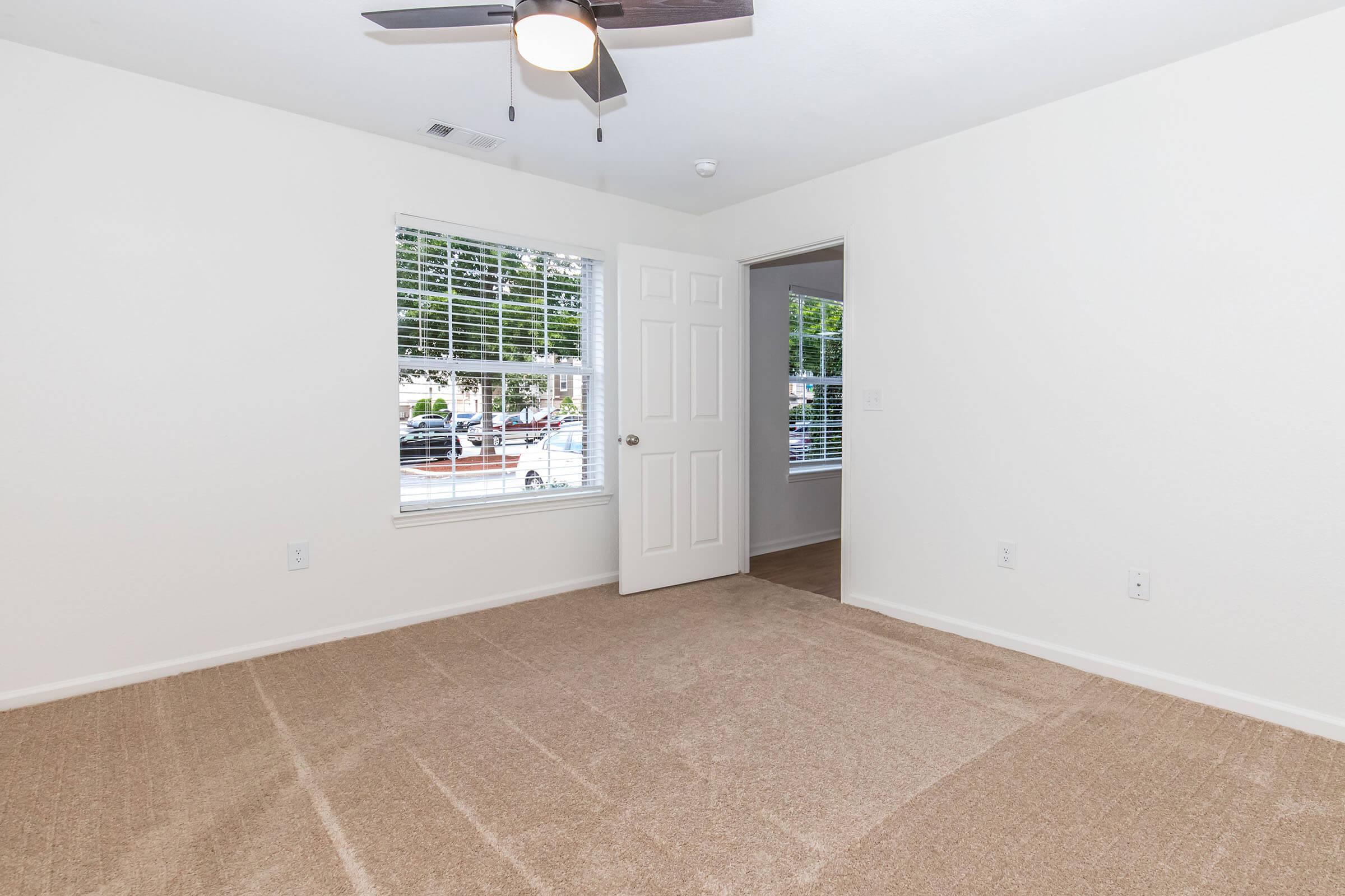 A spacious, empty room featuring light beige carpet, a ceiling fan, and large windows with white blinds. Natural light fills the space, and a door leads to another room. The walls are painted a neutral color, creating a fresh and clean environment, suitable for various uses.