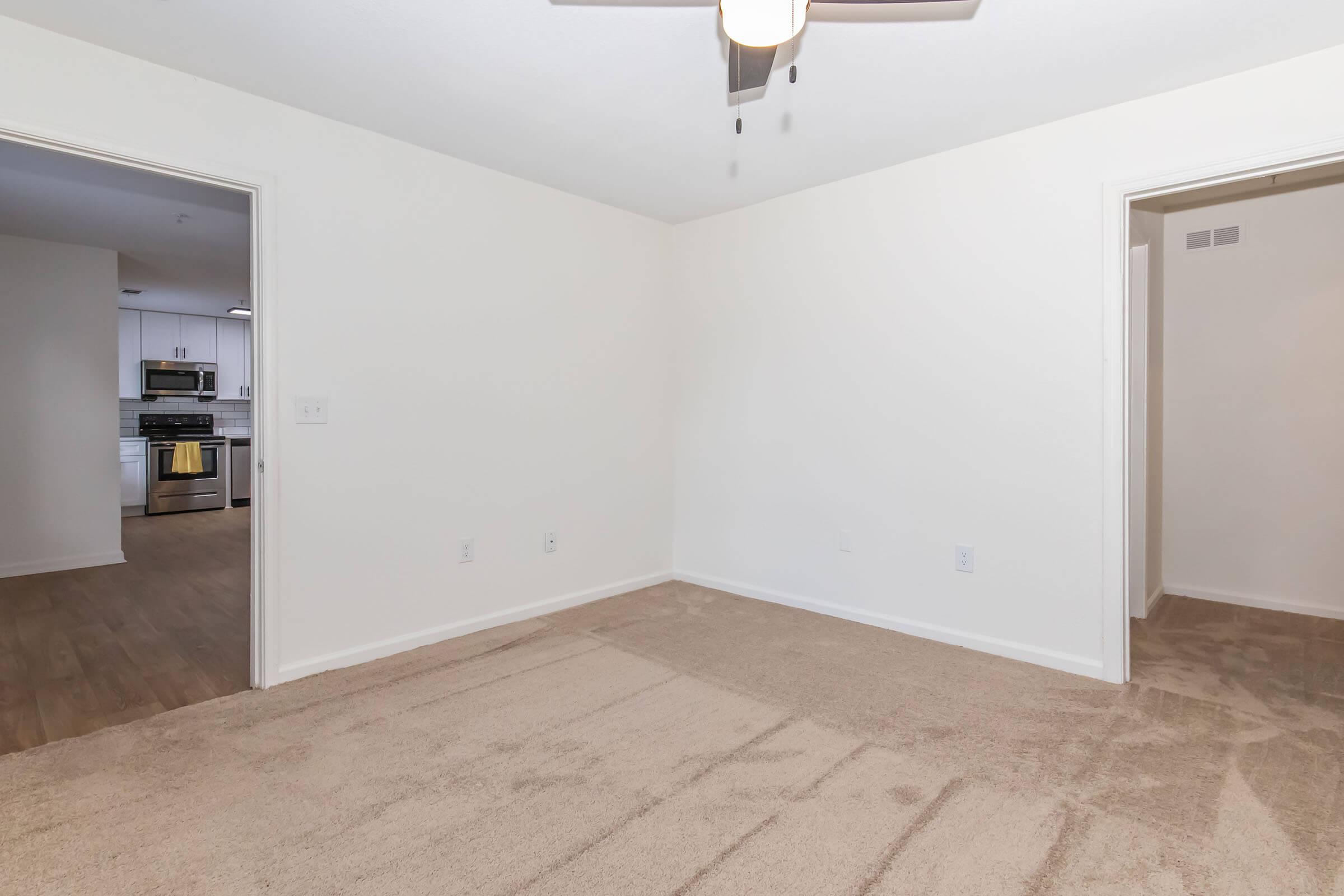 A vacant room with beige carpet and white walls, featuring a ceiling fan. There are two doorways leading to adjoining spaces. One doorway is partially visible, leading to a kitchen area with modern appliances, while the other doorway appears to lead to another room. The overall ambiance is bright and neutral.