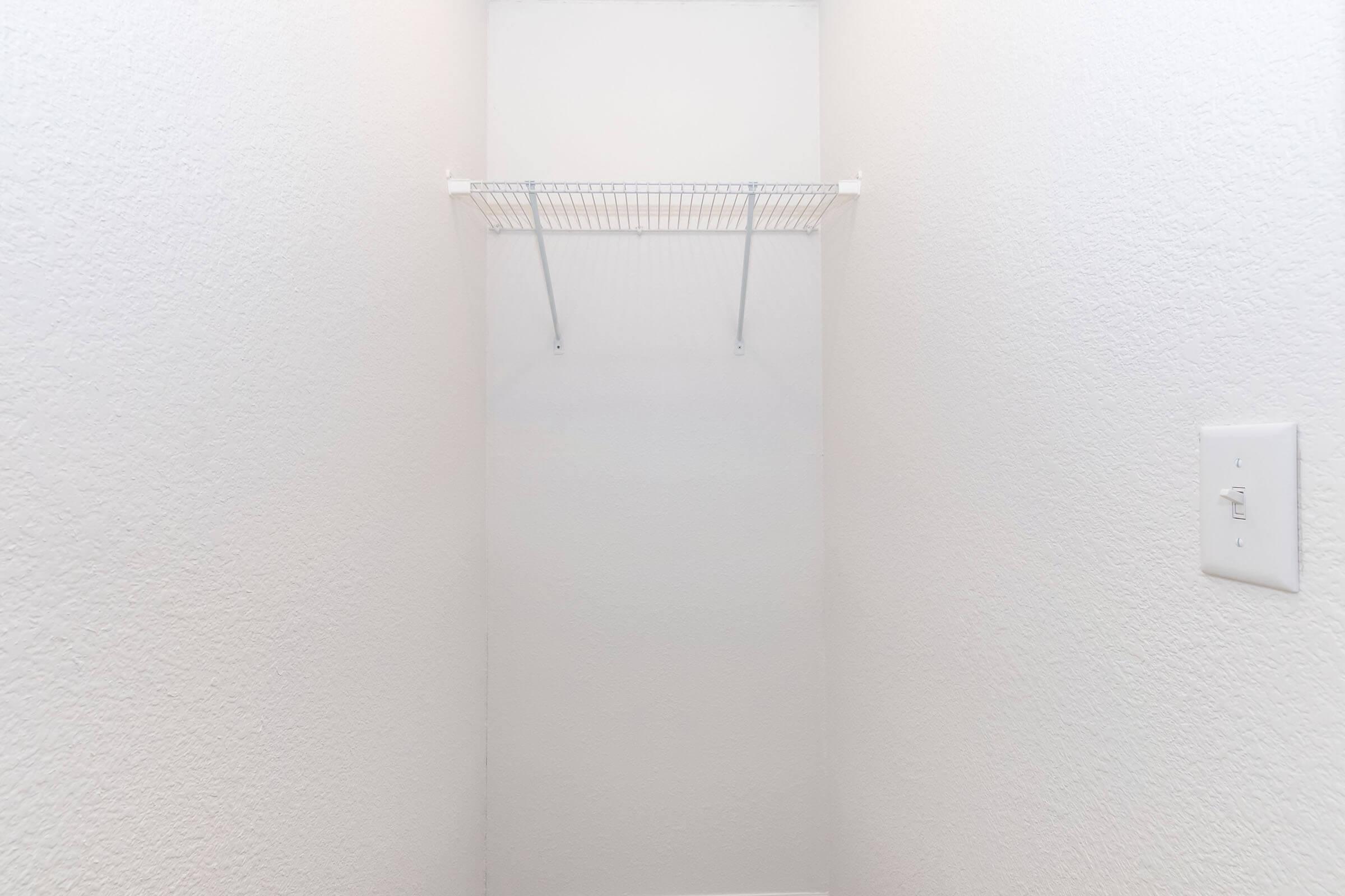 A minimalistic white closet with bare walls and a single wire shelf installed on the left side. The space is empty, emphasizing simplicity and openness, with a light switch to the right.