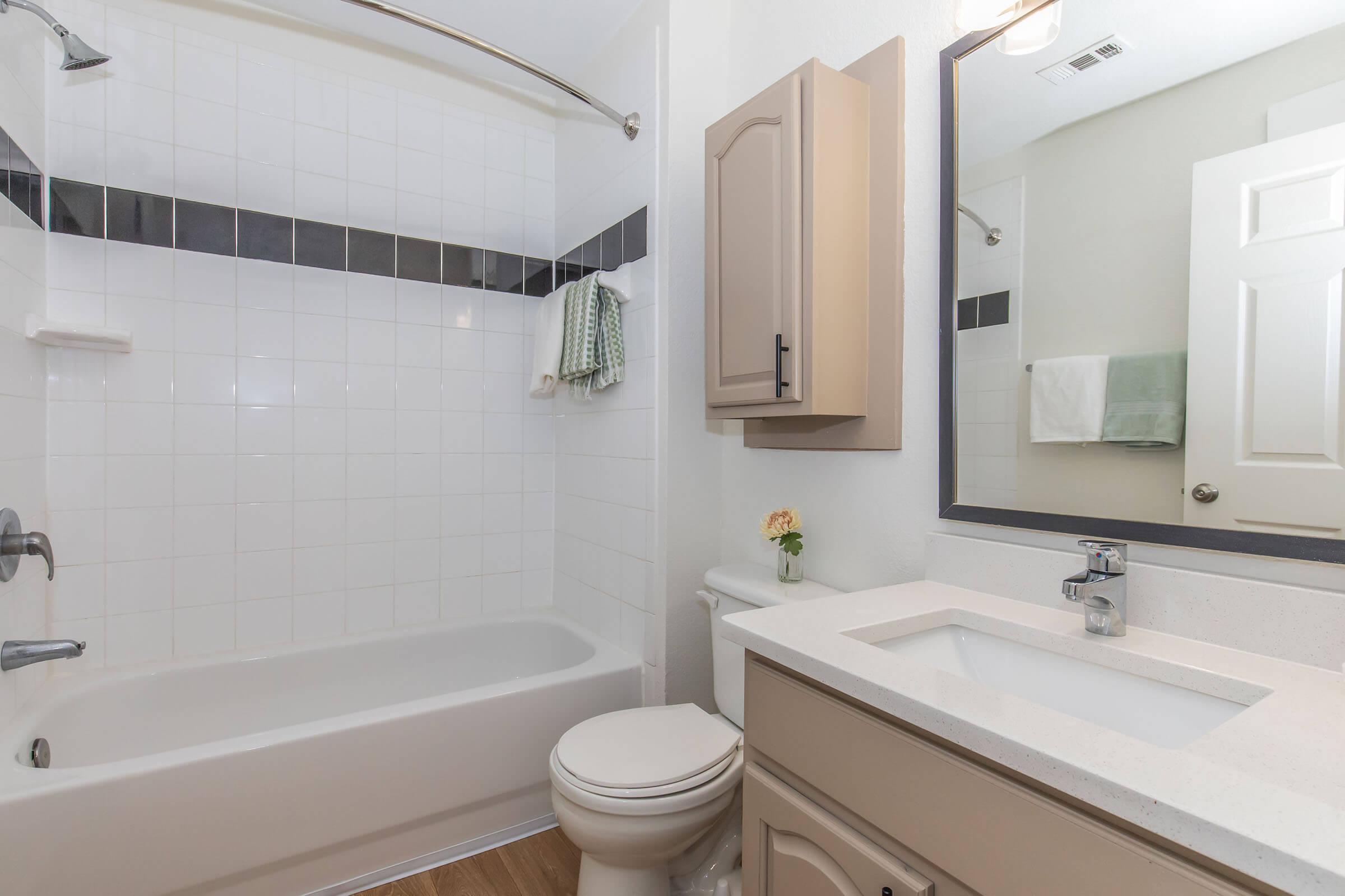 Modern bathroom featuring a bathtub with a shower rod, a wall-mounted mirror above a sink with a countertop, and beige cabinetry. Towel racks hold two neatly folded towels, and a small flower in a vase adds a decorative touch. The tiles are white with a dark accent strip.
