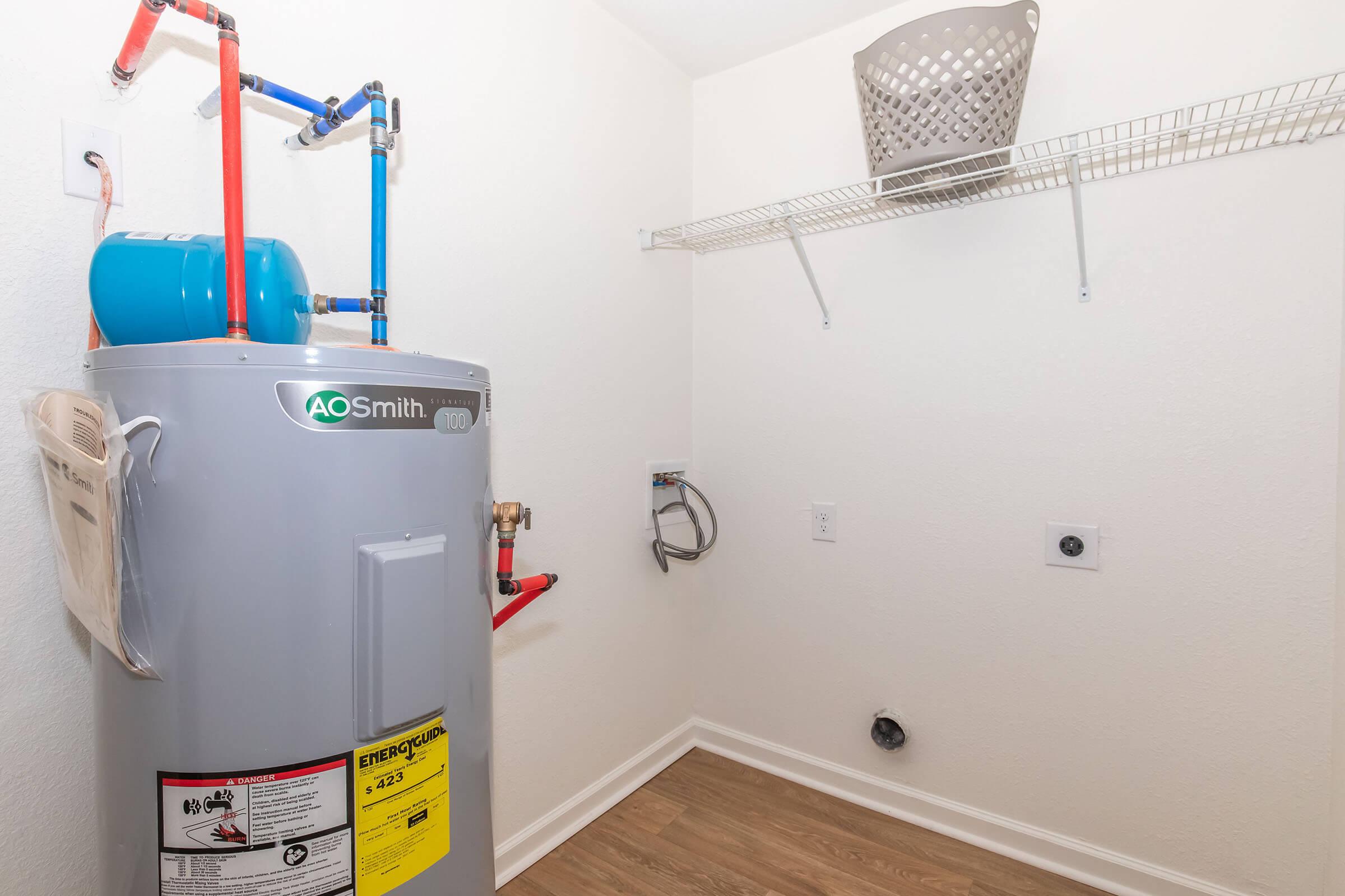 A laundry room featuring a gray AO Smith hot water heater, blue pipes, and a wire shelf. A white laundry basket sits on the shelf. The walls are painted white, and the floor has a wood-like finish. There is a wall outlet and plumbing connections visible in the space.