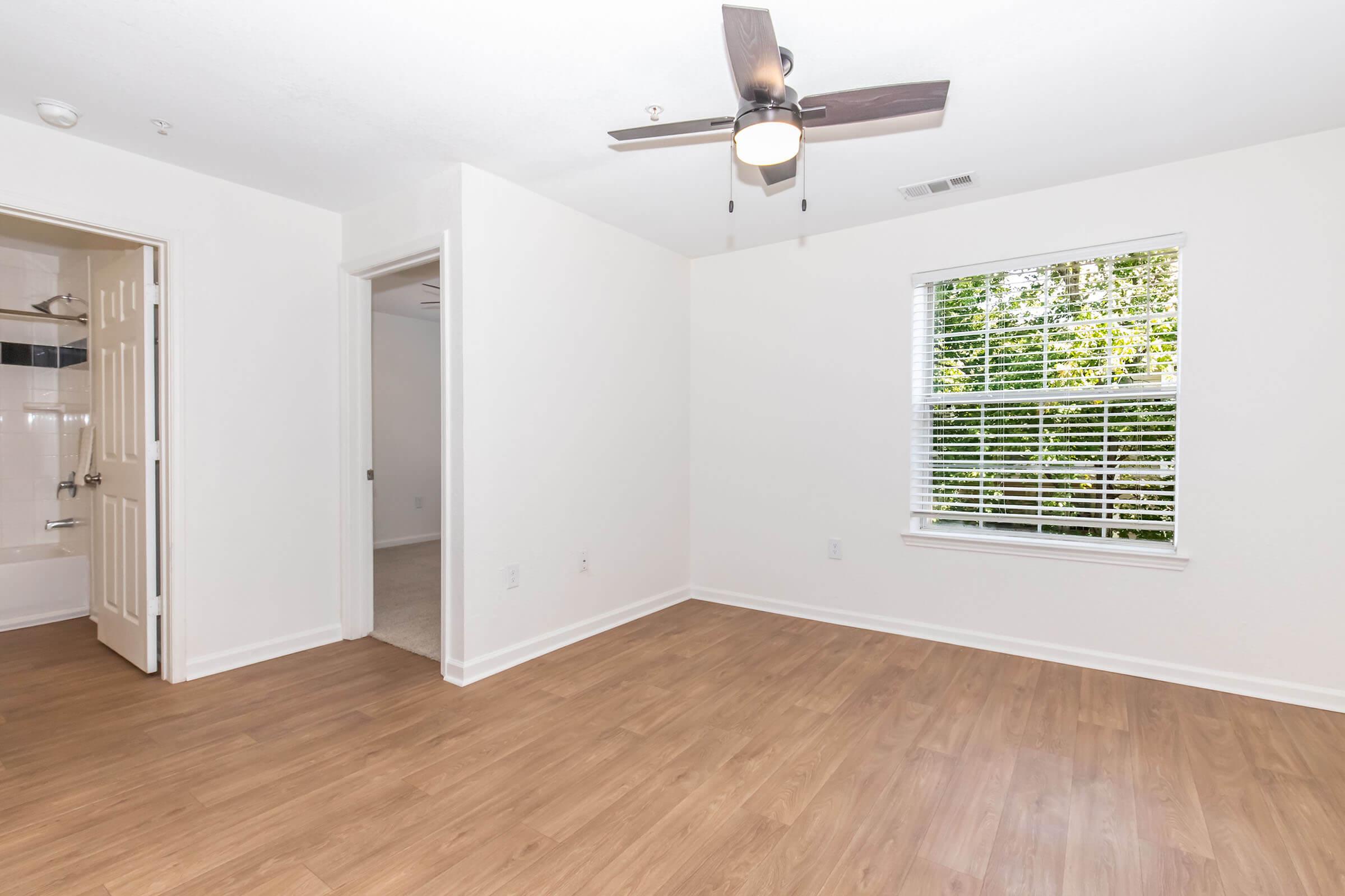 A clean and bright room featuring light wood-style flooring, white walls, and a ceiling fan. There is a window with blinds allowing natural light, and a doorway leading to another room. The layout is open, giving a spacious feel, and the room is ready for furniture placement.