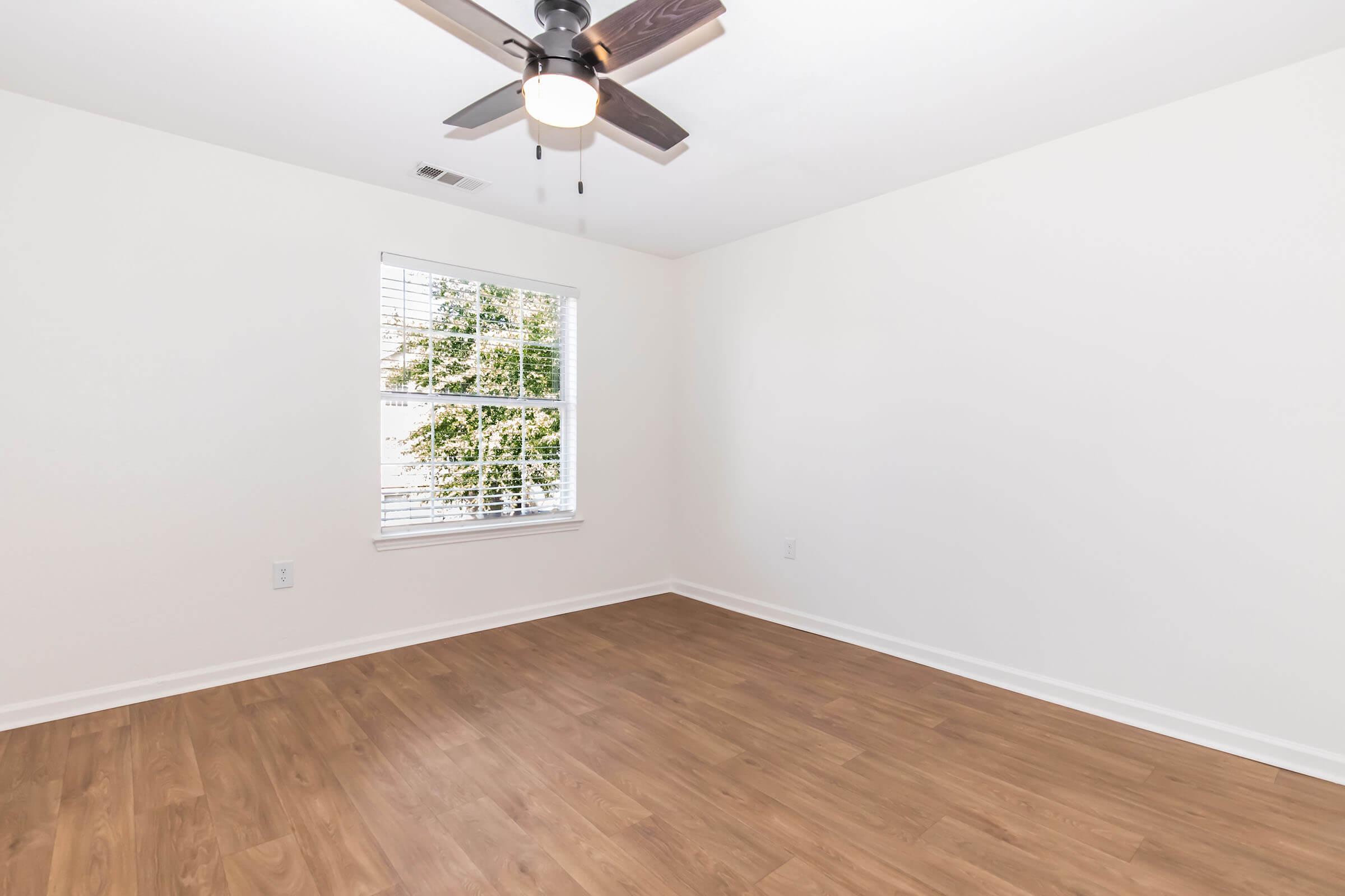 A bright, empty room featuring a light ceiling fan, wooden flooring, and a large window allowing natural light. The walls are painted white, and the space has a clean, modern feel, perfect for new furniture arrangement. The window offers a view of greenery outside.