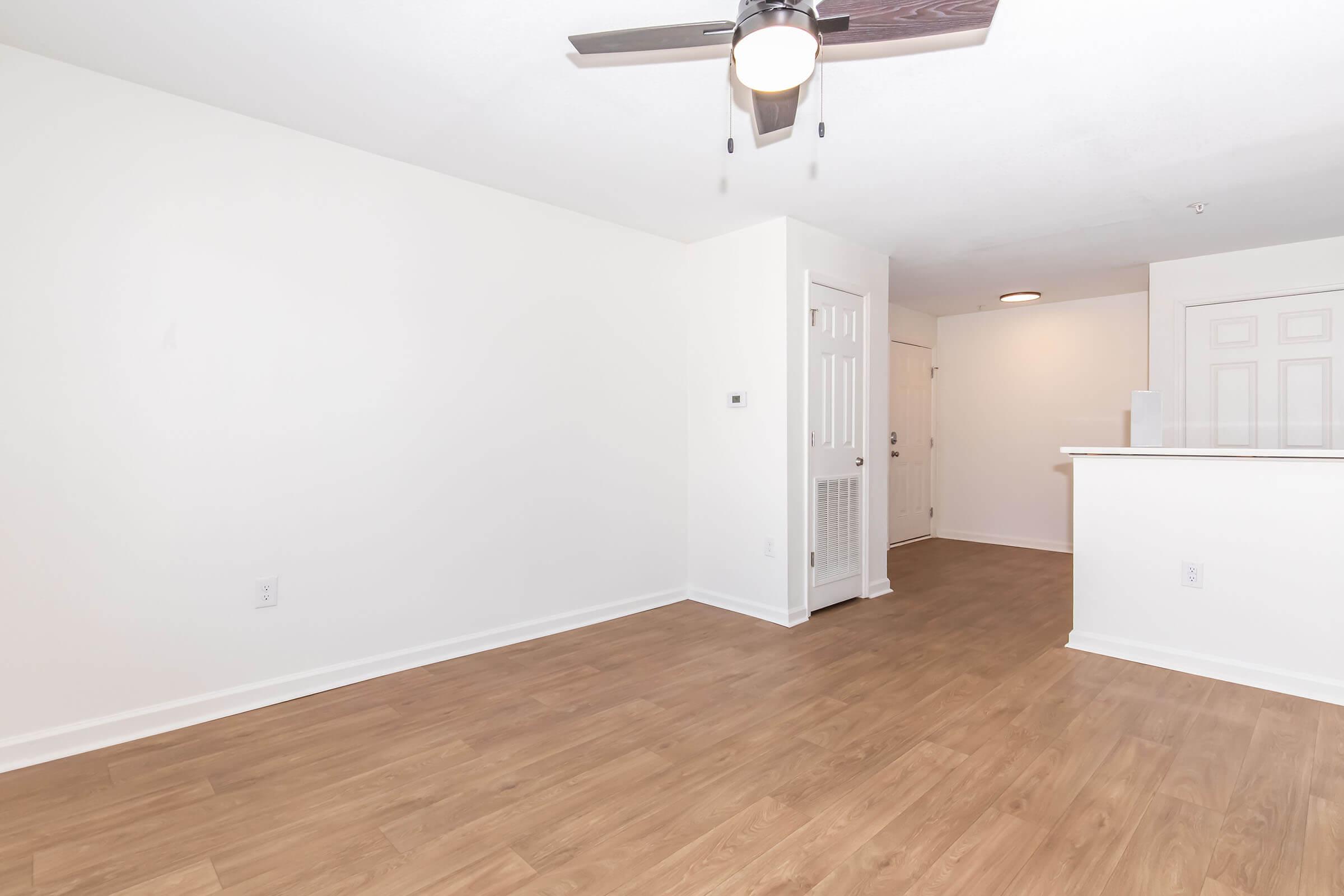 Spacious, empty living area with light-colored walls and wooden flooring. A ceiling fan is visible, along with a doorway leading to another room and an open kitchen area in the background. Natural light illuminates the space, creating a bright and inviting atmosphere.