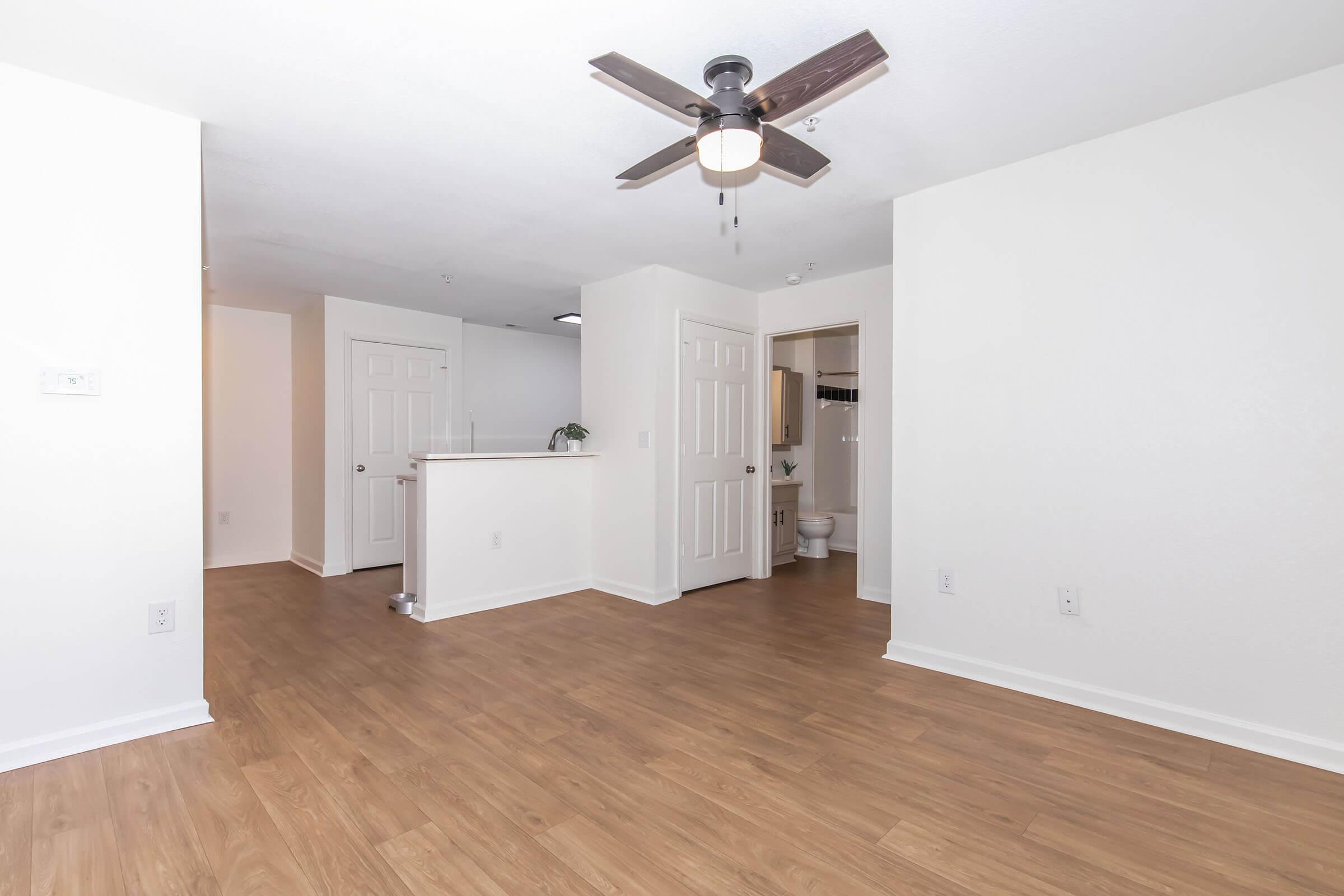 A bright, modern apartment interior featuring a spacious living area with light wood flooring, white walls, and a ceiling fan. There's an entryway on the left, and a hallway leads to a bathroom with an open door in the background. Natural light fills the space, creating an inviting atmosphere.