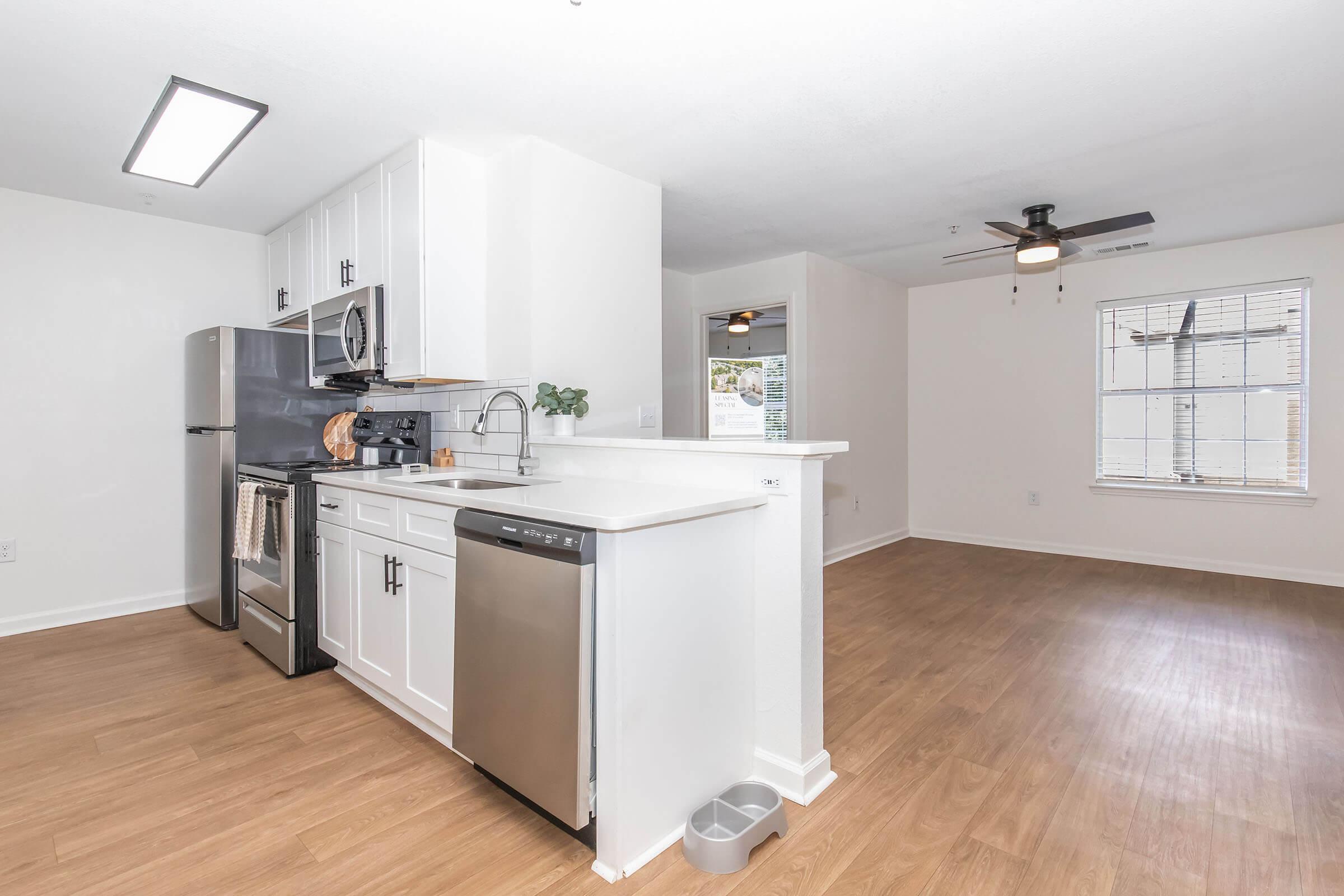 Modern kitchen with stainless steel appliances, including a refrigerator, microwave, and dishwasher. White cabinets and countertops create a clean look. The kitchen opens to a spacious living area with a ceiling fan and natural light streaming through large windows. Flooring is light wood.