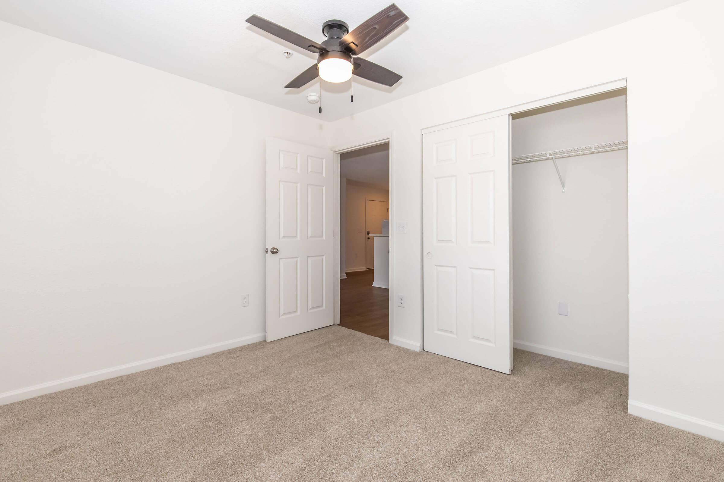 A clean, empty room featuring light beige carpet, two open closet doors with a small shelf, and a ceiling fan with a light fixture. The walls are painted white, and the space appears bright and well-maintained, suitable for various uses.