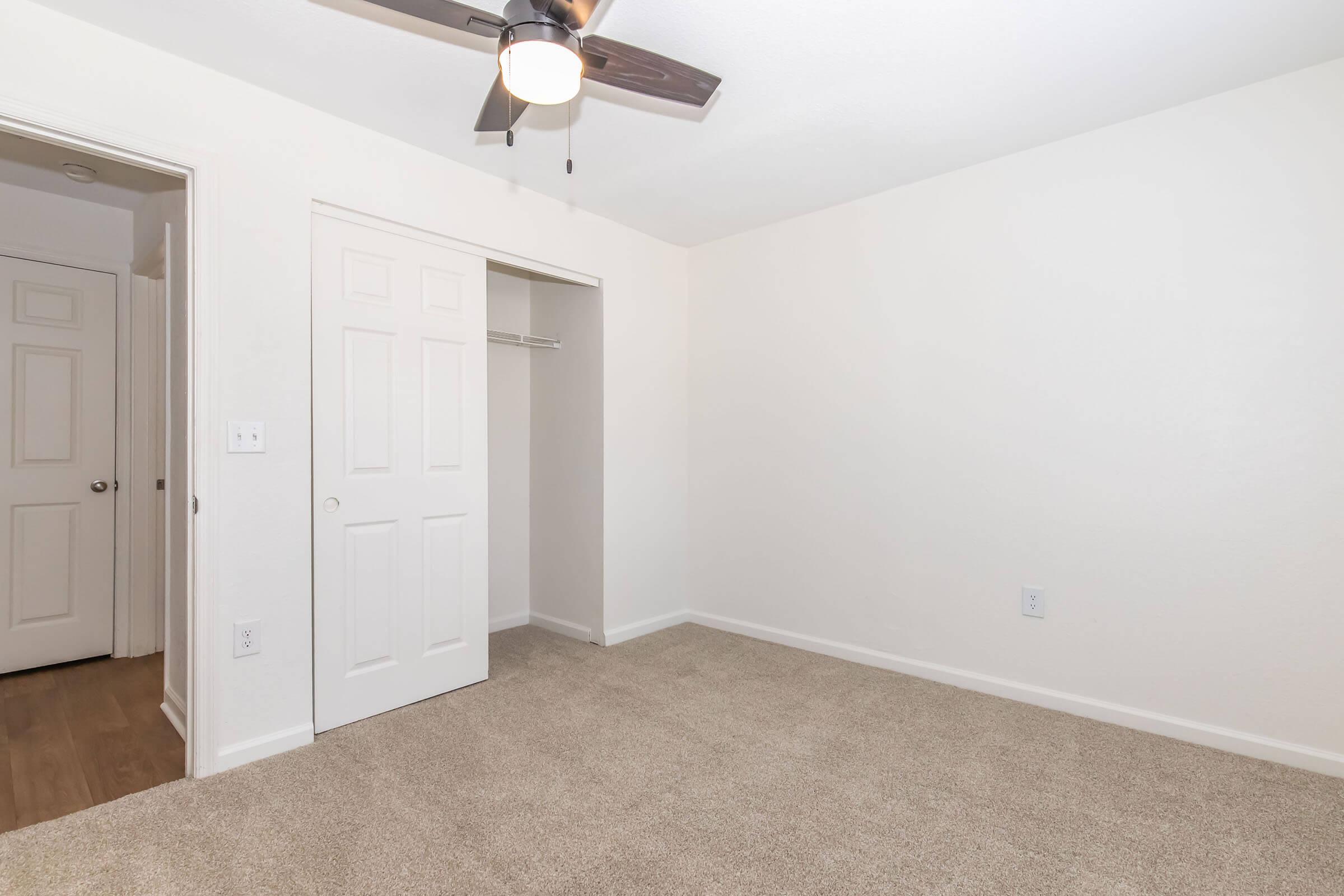 Empty room with light-colored walls and beige carpet. A ceiling fan is mounted on the ceiling, and there is a closet with a door partially open. Two doors are visible on the left, leading to adjacent spaces. The overall atmosphere is clean and minimalistic, suitable for a bedroom or an office.