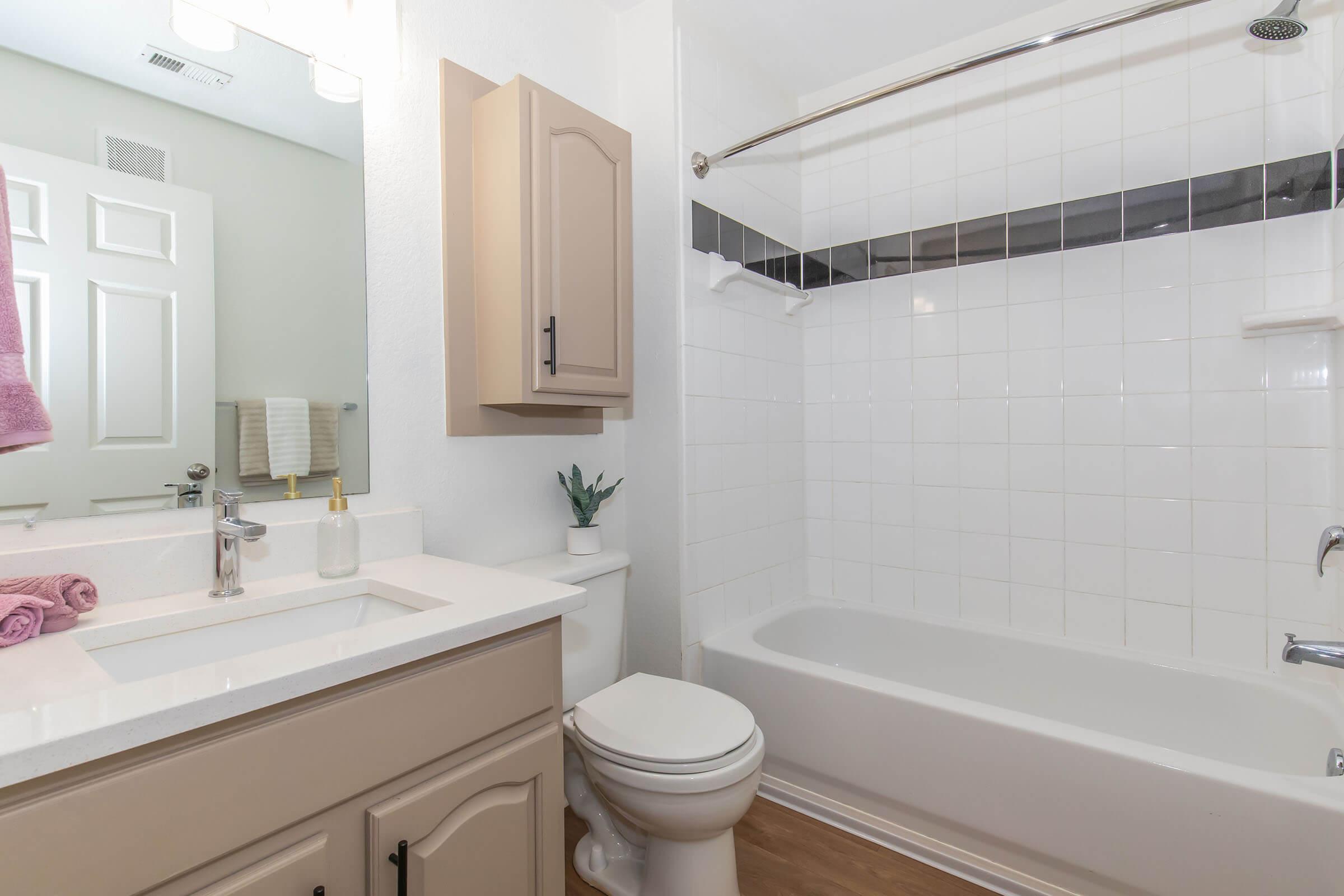 A clean and modern bathroom featuring a white bathtub, a sink with a mirror above, and a beige cabinet. There are towels hanging, a small plant on the counter, and neutral-toned flooring. The space is well-lit with a bright ceiling light.