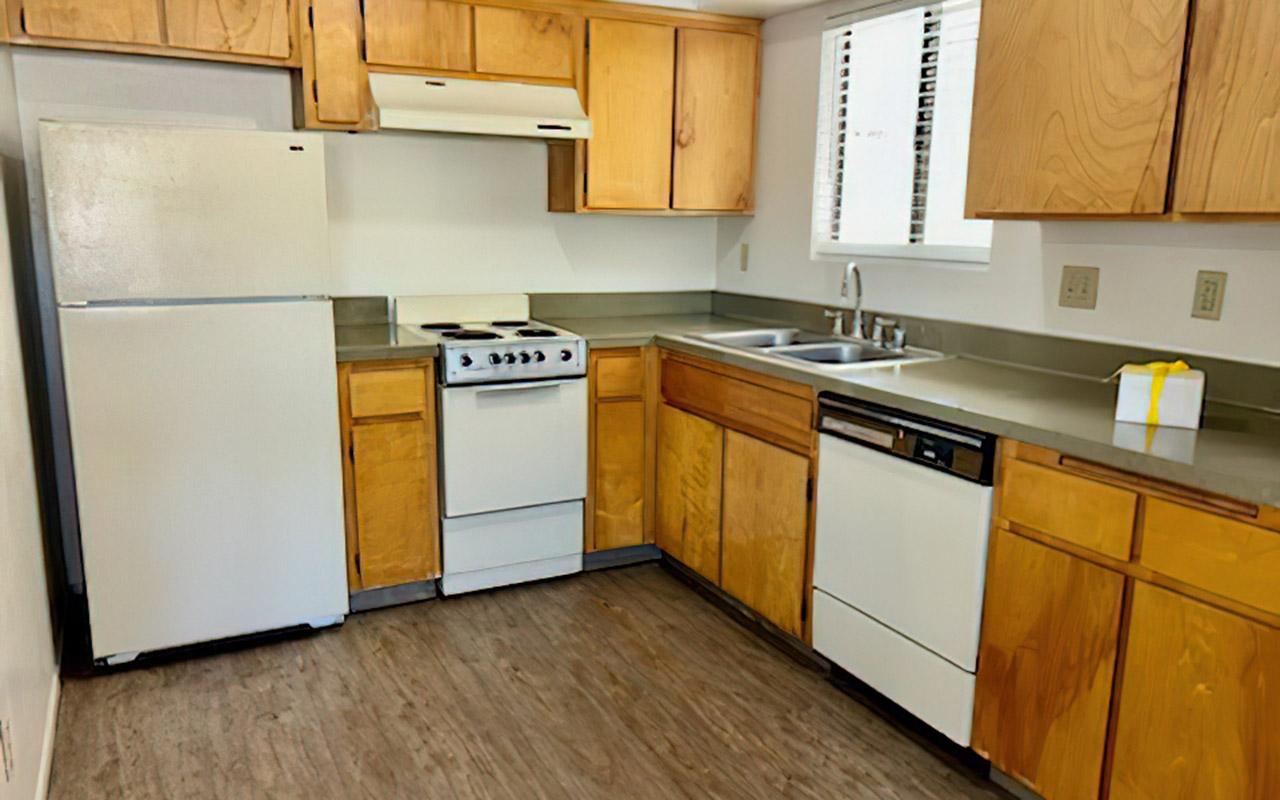 A modern kitchen featuring wooden cabinetry, a white refrigerator, a gas stove with four burners, a sink with a window above, and a dishwasher. The countertop is gray, and the flooring is wood-like. There's a small white box on the countertop, adding a touch of color to the space.