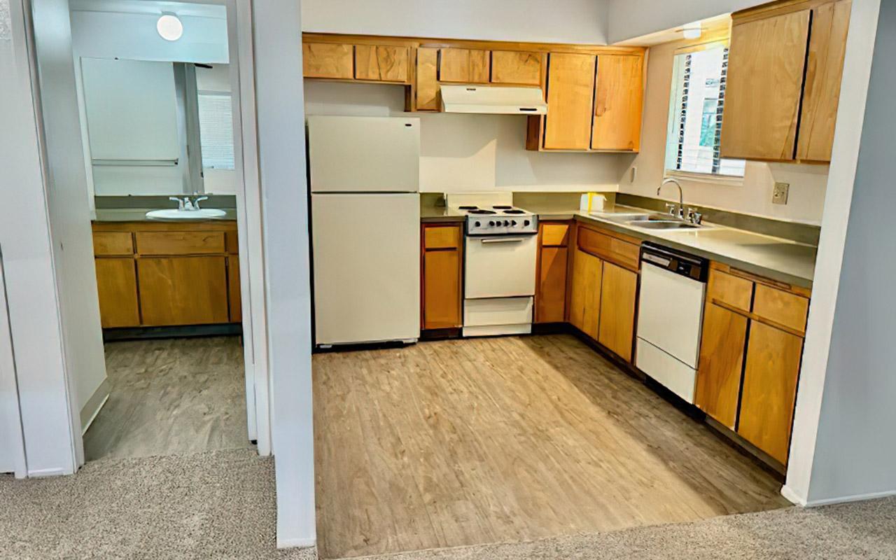 A bright kitchen featuring wooden cabinets, a white refrigerator, a gas stove, and a dishwasher. The countertop is light-colored, and there's a window providing natural light. The adjoining area has a sink and additional space, with simple flooring and neutral-colored walls, creating a cozy and functional atmosphere.