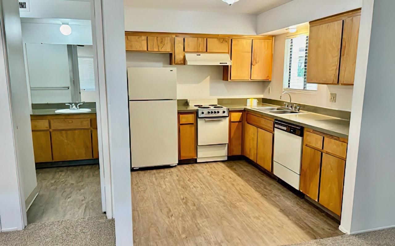 A spacious kitchen featuring wooden cabinets, a refrigerator, stove, and dishwasher. The countertop is complemented by a window letting in natural light. The layout includes an adjoining room visible in the foreground, showcasing carpeted flooring and an entrance to a bathroom area.