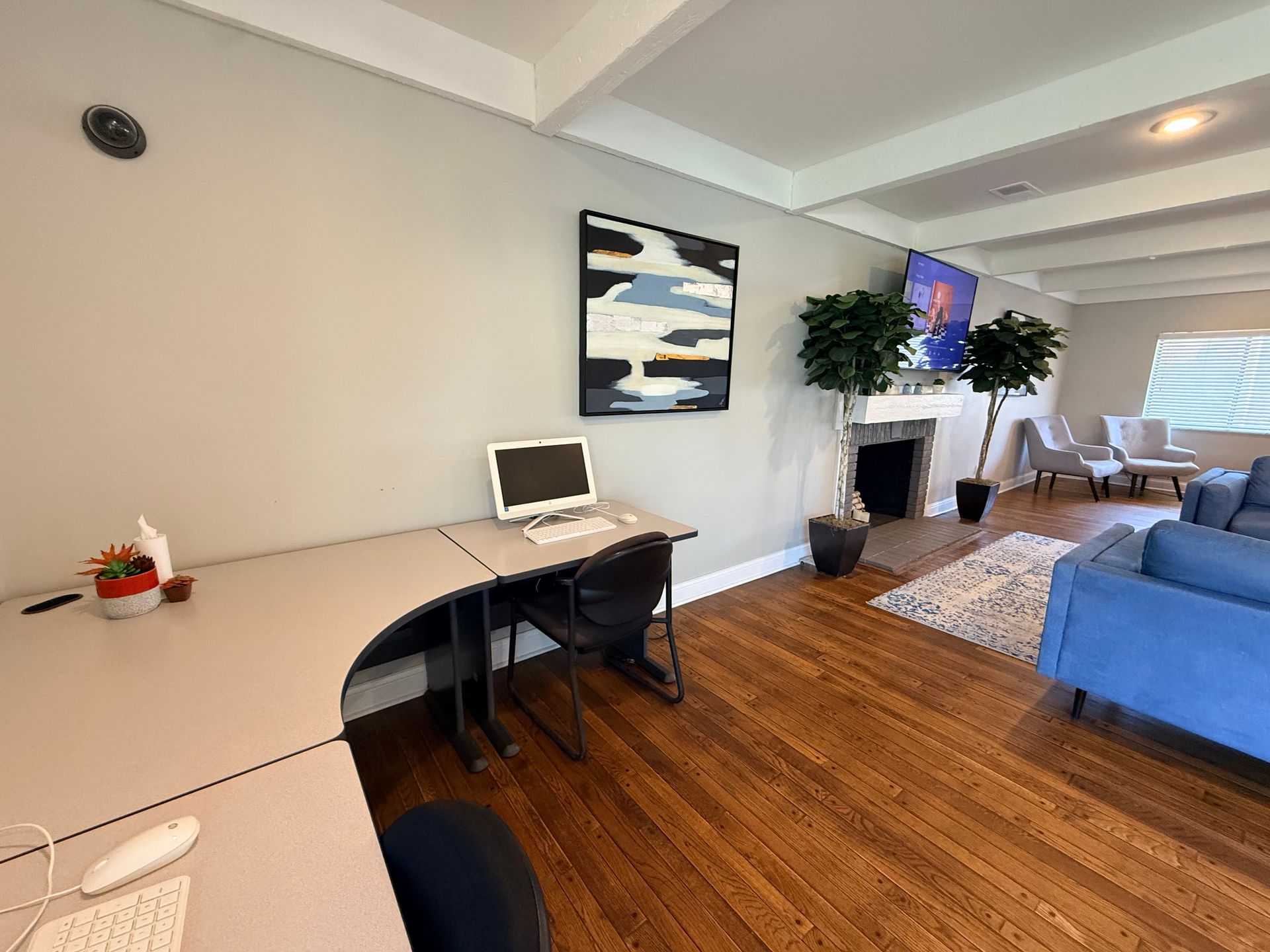 An office space featuring a desk with a computer, mouse, and a small plant. To the right, a cozy living area with a blue couch and two chairs, a fireplace, and a television mounted on the wall. The room has light-colored walls and wooden flooring, with a piece of modern art hanging above the desk.
