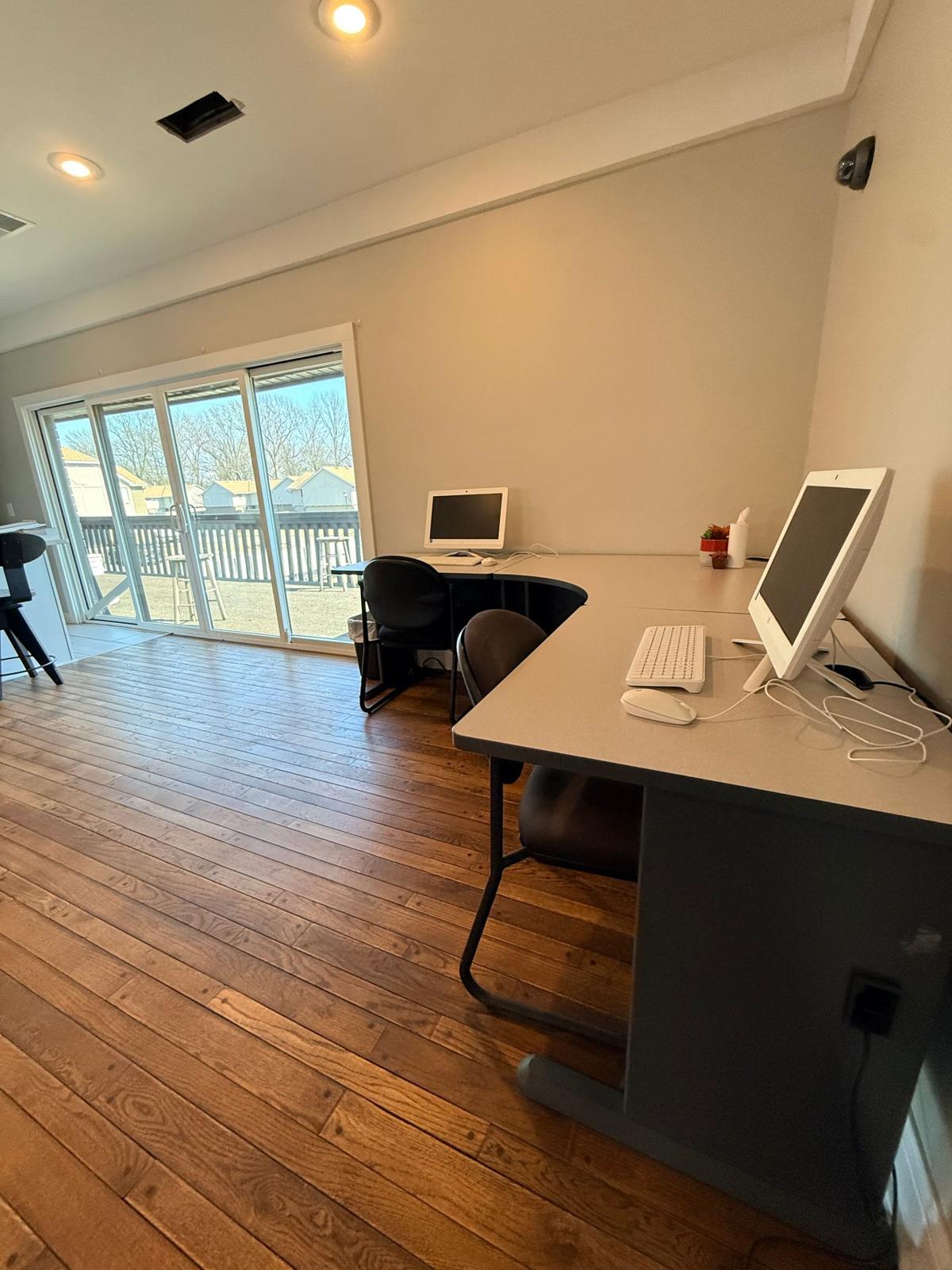 A bright, spacious room featuring two computer desks with monitors and keyboards. The hardwood floor adds warmth to the space, while large windows let in natural light, offering a view of the outdoors. The minimalist design creates a clean, functional atmosphere suitable for work or study.