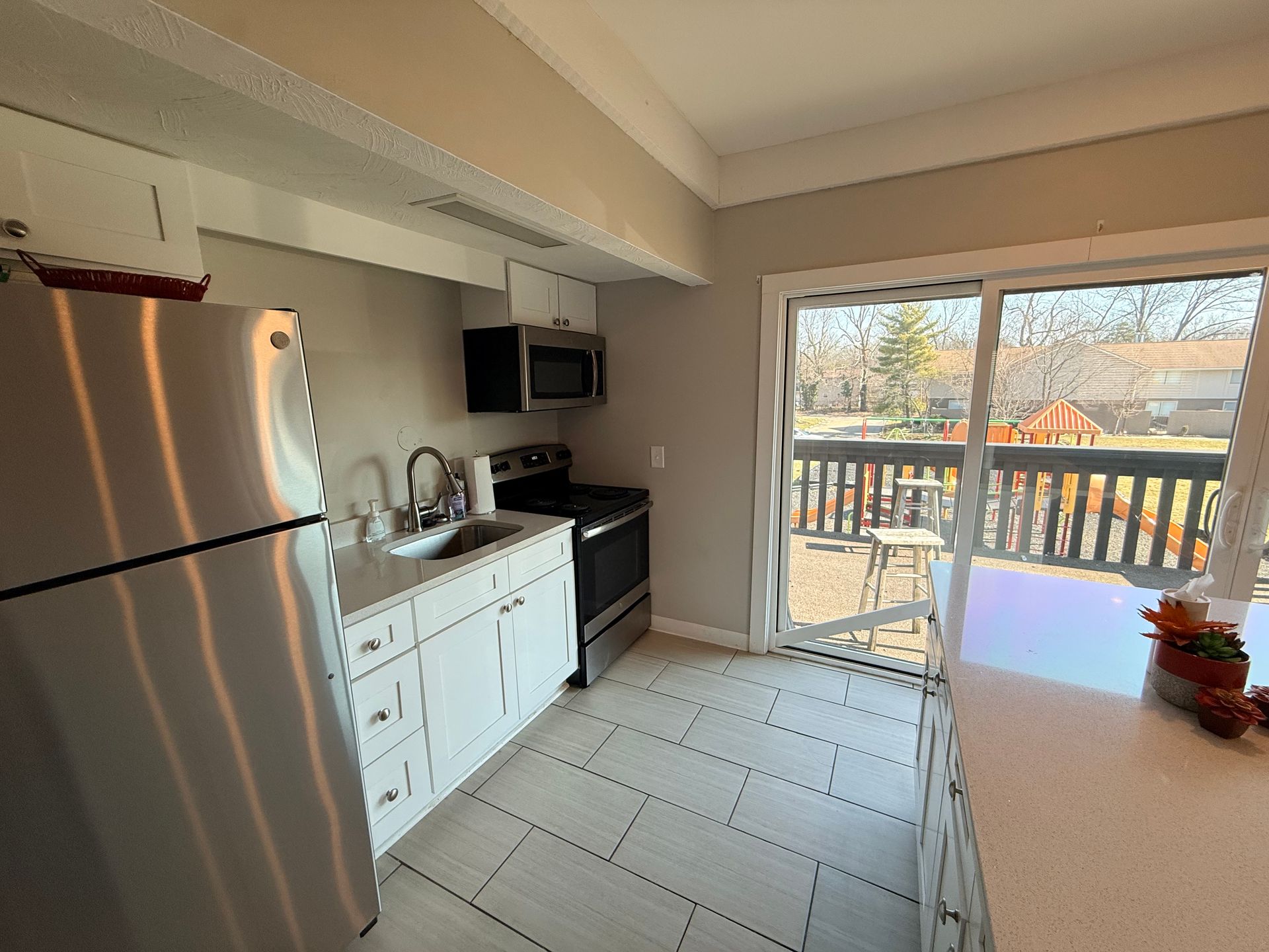 A bright kitchen featuring stainless steel appliances, including a refrigerator and an oven, complemented by white cabinetry. There is a countertop with a sink, and a sliding door leading to a deck area outside, allowing natural light to fill the space. The flooring is tiled in light colors.