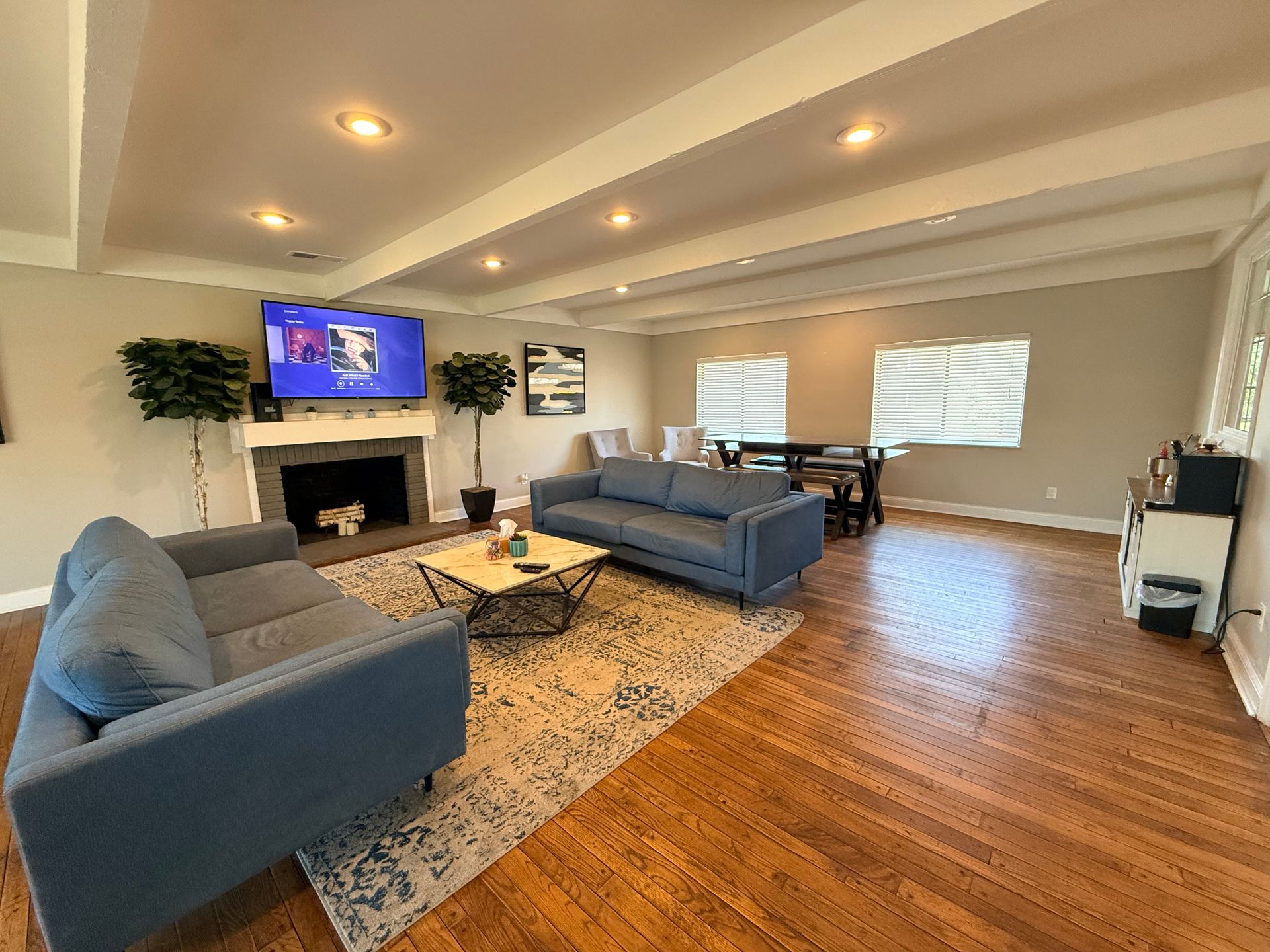 A spacious living room with two blue sofas facing a coffee table on a patterned rug. A flat-screen TV is mounted above a fireplace. In the background, there is a dining area with a table and chairs. Windows on either side allow natural light to illuminate the room, which features hardwood flooring and neutral walls.