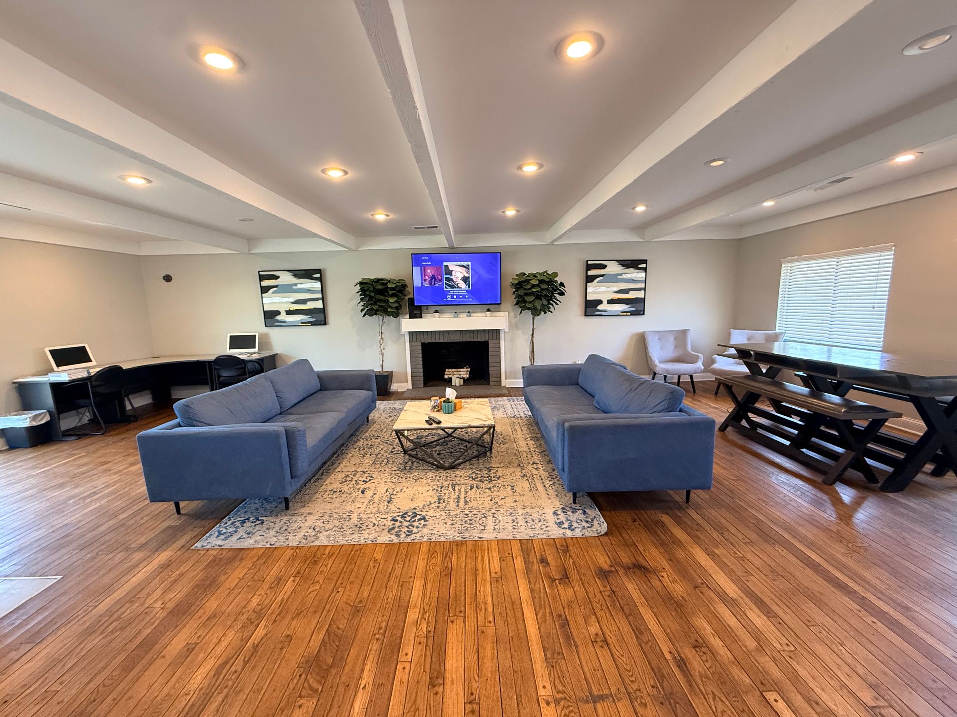 A spacious living room featuring two blue sofas facing a coffee table, with a television mounted on the wall. There are two plants flanking the fireplace, a dining table to the right, and two workstations with computers on the left. Wooden floors add warmth to the modern decor.