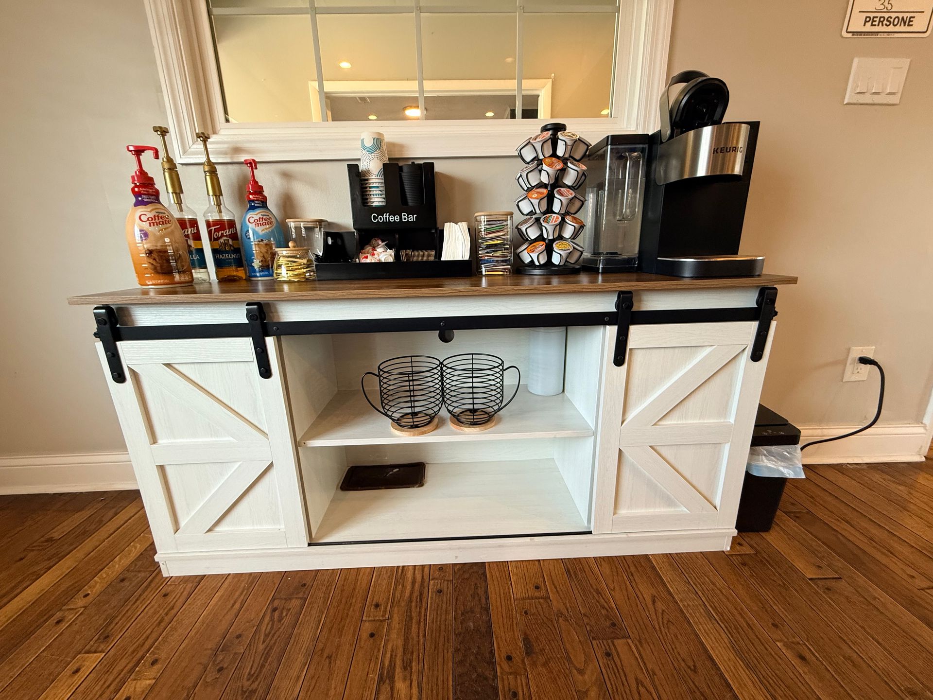 A coffee bar station featuring a wooden countertop, coffee machine, various coffee syrups, a carousel of coffee pods, and storage bins. The unit has a rustic design with sliding barn doors, set against a neutral wall, with a mirror reflecting the space, and a trash can visible to the side.
