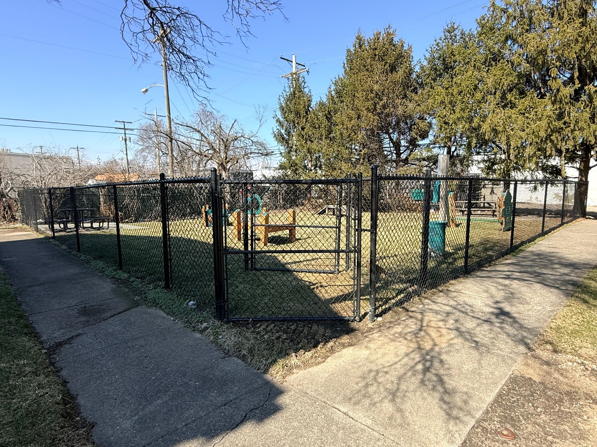 A fenced playground area featuring wooden play structures on green grass, surrounded by trees and utility poles. Two adjacent sidewalks lead to the entrance of the playground, providing access from either side. The sky is clear and blue, indicating a sunny day.
