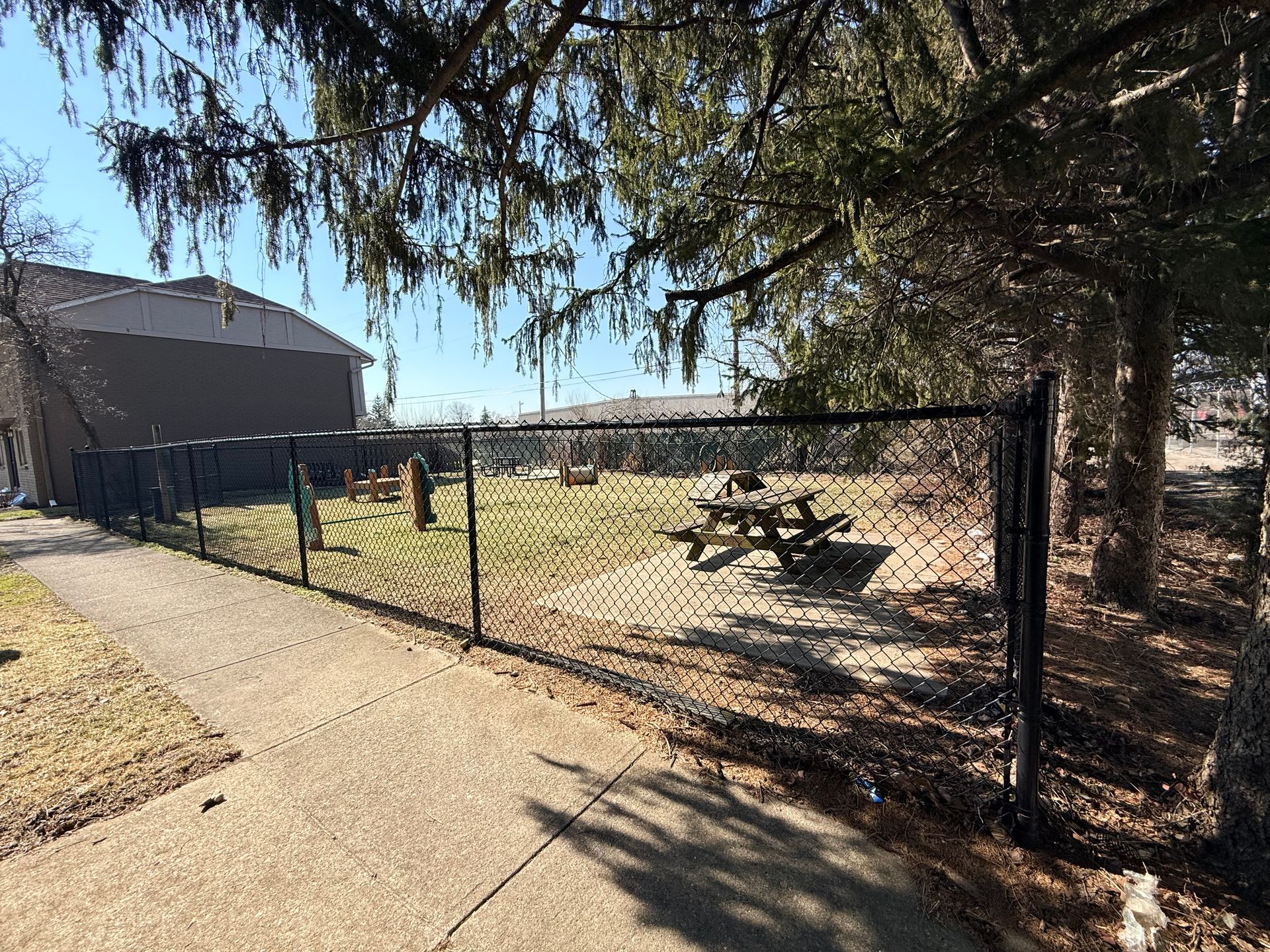 A fenced area with a picnic table and benches on a grassy section, surrounded by trees. The scene shows a clear blue sky and a sidewalk leading up to the fence. In the background, there are buildings visible, indicating an industrial or community setting.