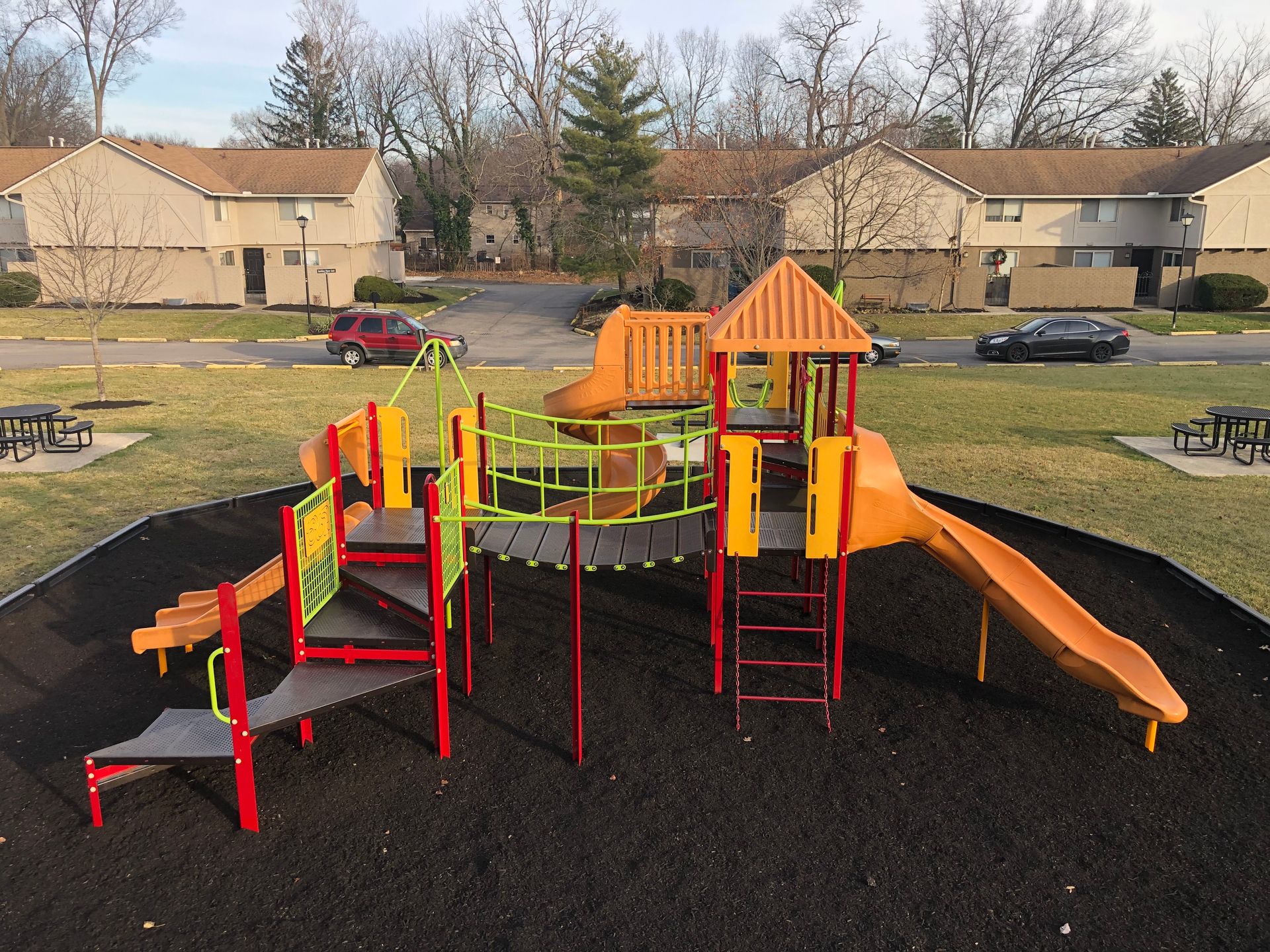 A vibrant playground featuring a variety of play structures, including climbing equipment, slides, and a bridge, set in a grassy area. Surrounding the playground are picnic tables and residential buildings in the background, creating a family-friendly environment.