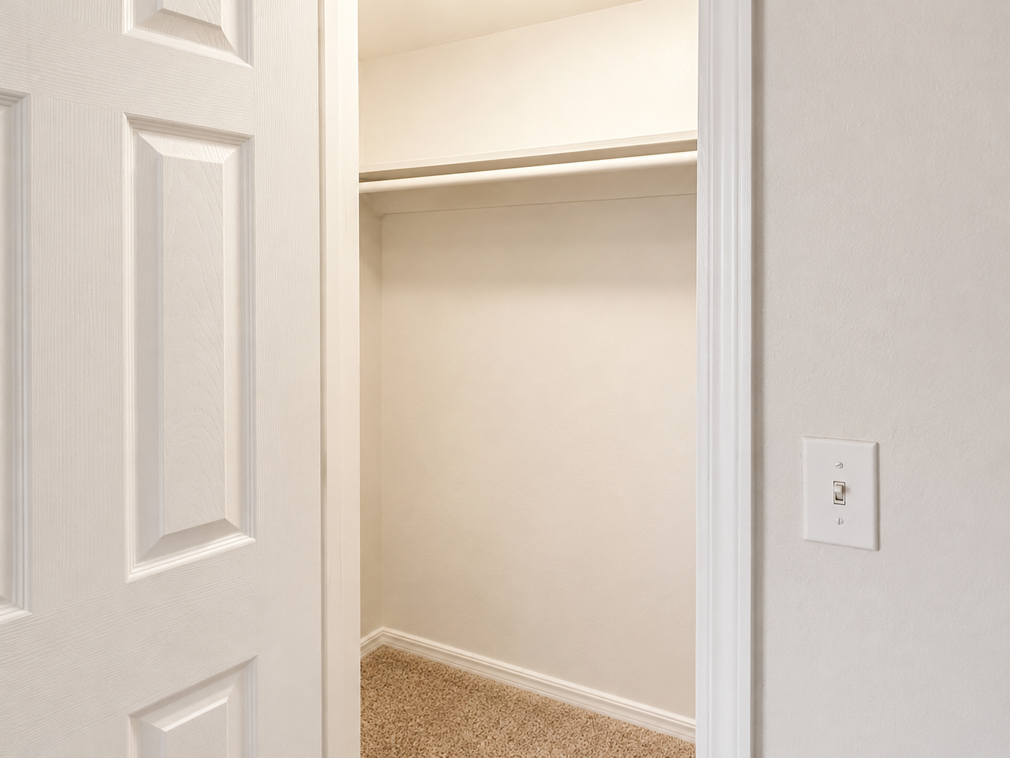 A clean, empty closet with a light-colored interior. The door is partially open, revealing a spacious, uncluttered shelf and beige carpet flooring. The wall is smooth with a subtle texture, and there is a light switch on the adjacent wall.