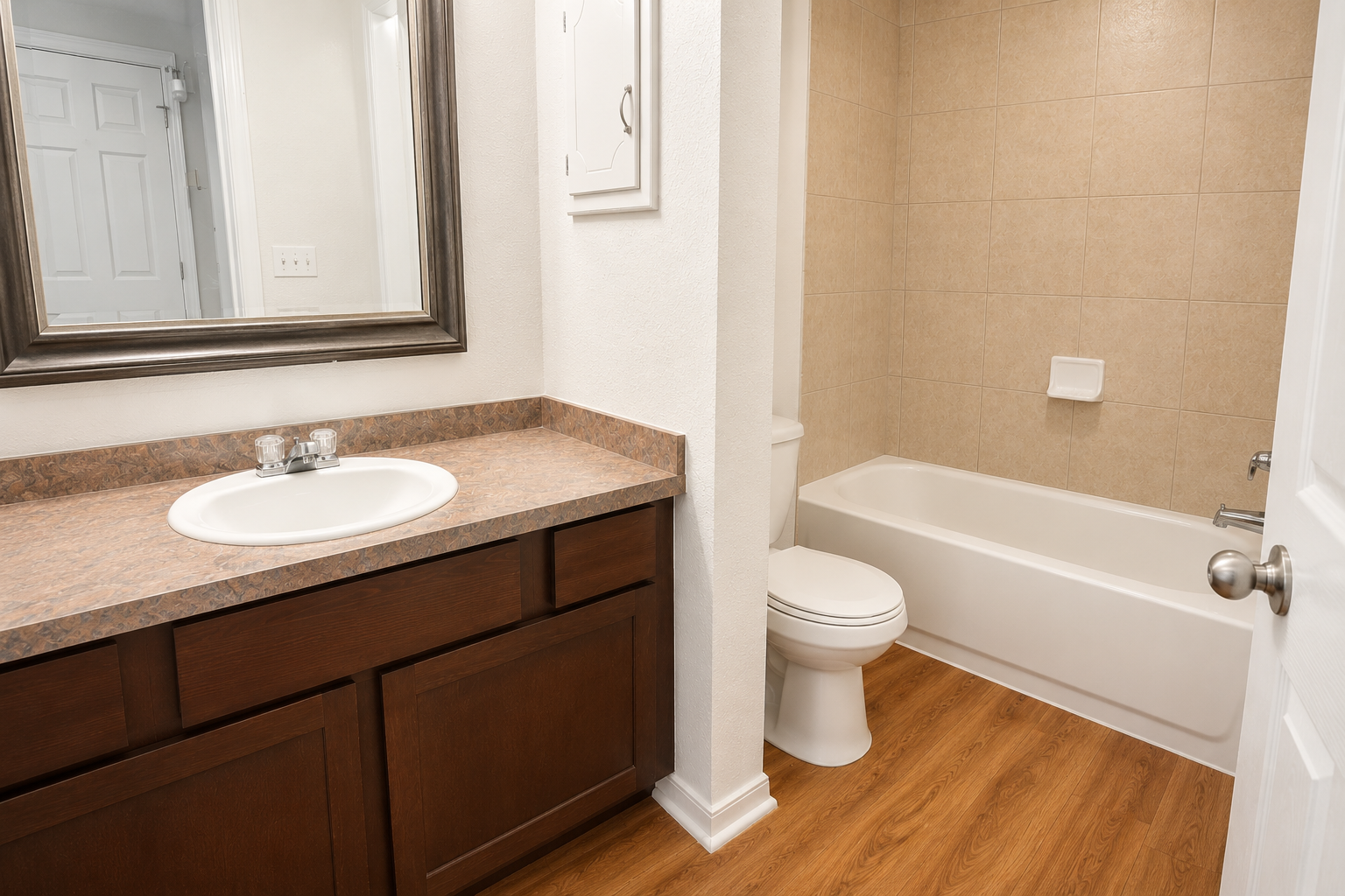 A clean and modern bathroom featuring a white bathtub, toilet, and a sink with a brown countertop. The walls are tiled in a light beige color, and there is a large mirror above the sink. The floor has a wood-like finish, and a door leads to an adjacent room. Natural light brightens the space.