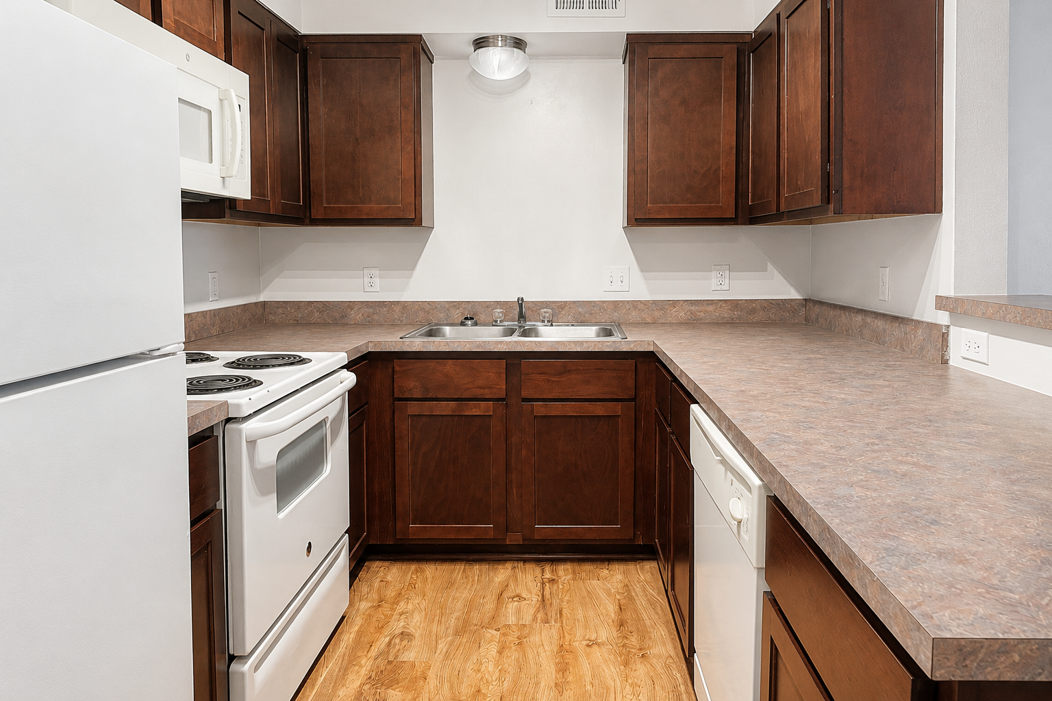 A modern kitchen with dark wooden cabinets and a light countertop. The appliances include a white refrigerator, a white stove with an oven, a sink with dual faucets, and a dishwasher. The floor is a warm wood-like laminate, creating a comfortable and inviting atmosphere.