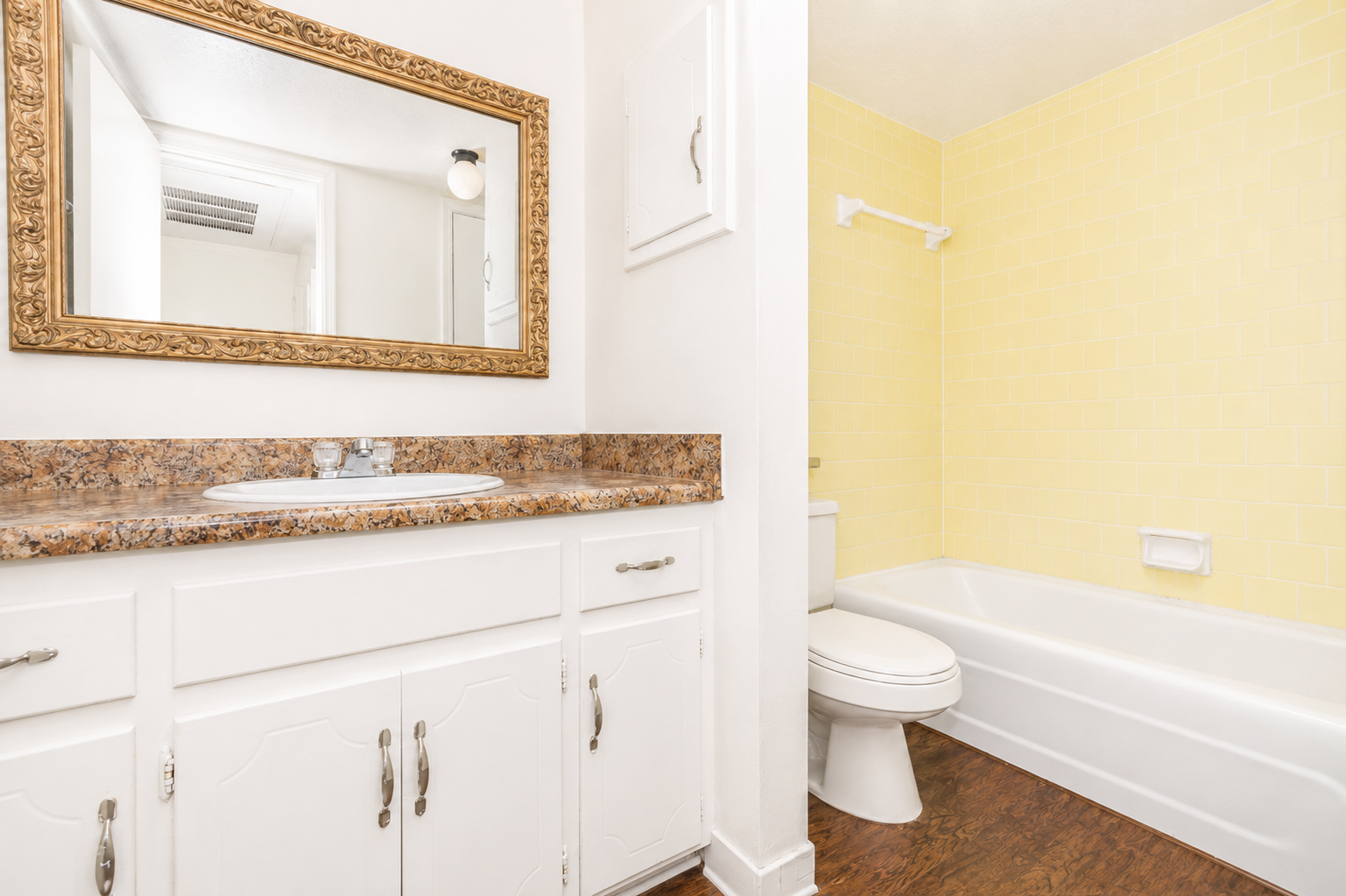 A bright bathroom featuring a white sink with a granite countertop and a large ornate mirror. The walls are painted yellow, and the space includes a bathtub, toilet, and a small cabinet above the sink. Soft lighting from a ceiling fixture enhances the warm atmosphere.