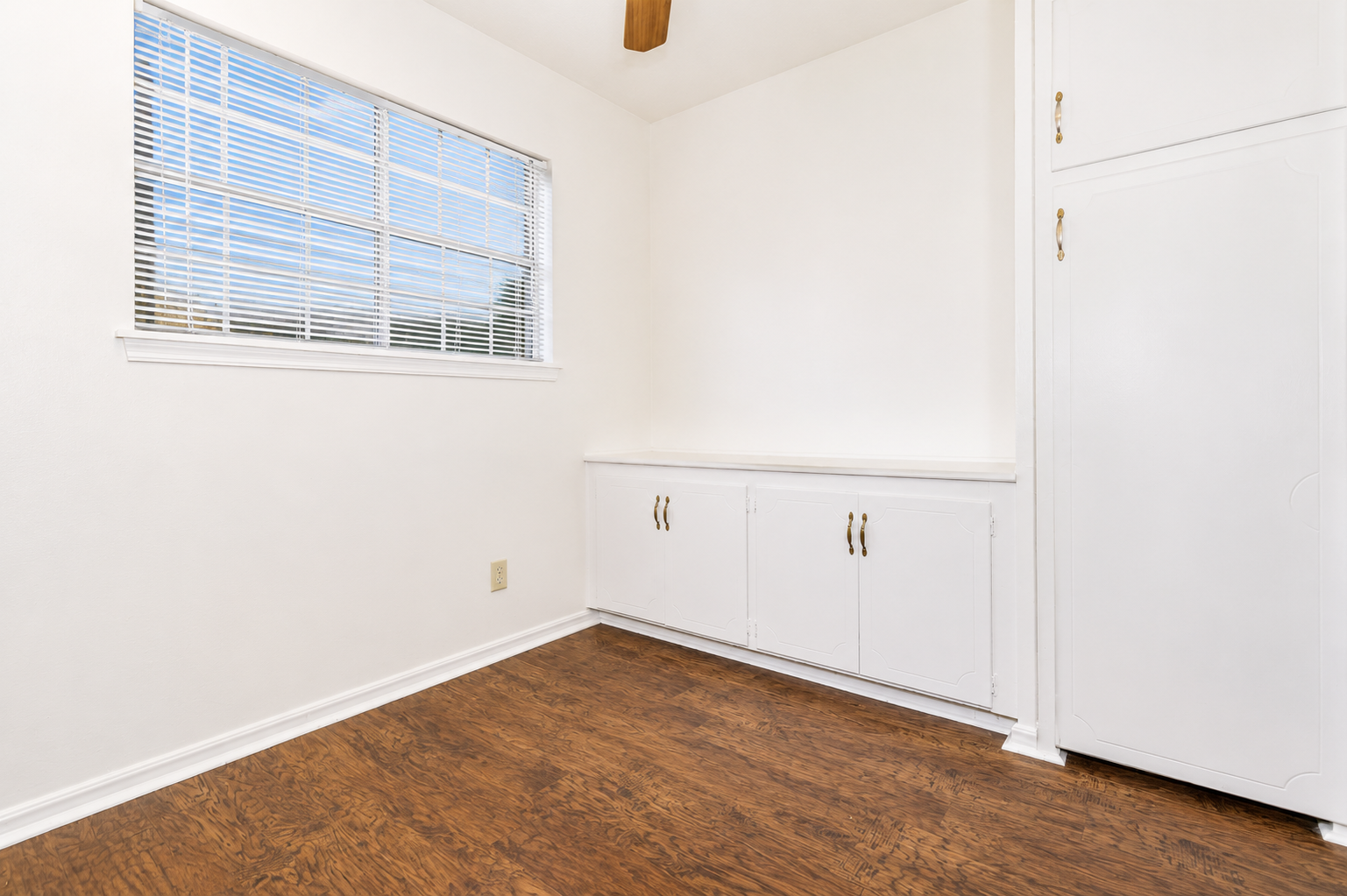 A bright, empty room featuring a large window with white blinds, a smooth light-colored wall, and a built-in white cabinet with gold handles. The floor is made of dark wooden planks, creating a contrast with the light walls. A ceiling fan is visible above, adding to the room's airy feel.