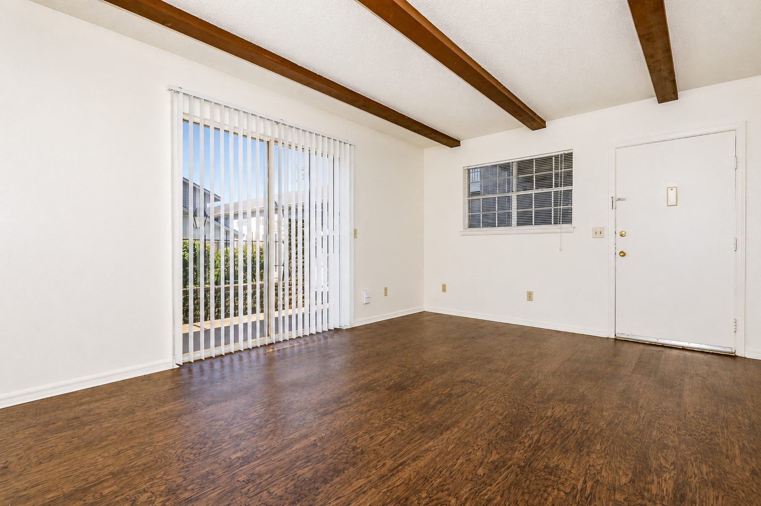 A spacious, empty room with wooden beam ceilings, featuring a sliding glass door that leads to the outside, a small window, and a front door with a keypad entry. The floor is a light wood laminate, and the walls are painted white, creating a bright and airy atmosphere.
